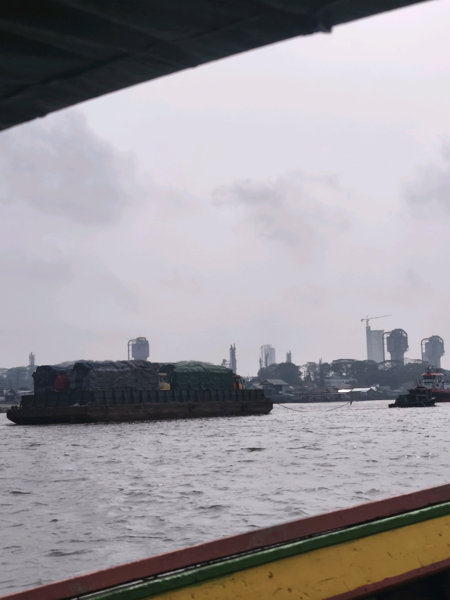 The image depicts a view from a boat on a wide river, likely the Musi River in Palembang, Indonesia, given the presence of industrial structures in the background. The sky is overcast, creating a muted light.
In the foreground, the edge of the viewing boat is visible, with a red, green, and yellow painted railing.
The main subject of the image is a large barge loaded with goods covered by dark tarpaulins. The barge is positioned in the middle ground and appears to be in motion. A small yellow cabin or structure is visible on the deck of the barge.
In the background, the cityscape of Palembang is discernible. Several tall buildings, some appearing to be part of industrial complexes or high-rise residential/commercial structures, dominate the skyline. A construction crane is visible near one of the taller buildings. There are also indications of smaller buildings and trees along the riverbank.
A smaller tugboat is also visible in the mid-right of the frame, seemingly accompanying the barge or engaged in nearby activity on the river. The water is choppy, indicating a steady current or wind. The overall scene suggests a busy industrial or shipping area.