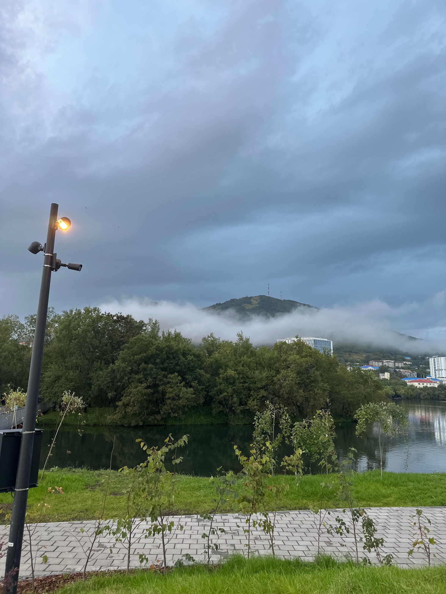 The image depicts a park area with a paved walkway in the foreground, leading to a body of water. A lamppost with an illuminated light and two cameras is positioned to the left of the frame. Beyond the water, dense green trees form a natural barrier, with a cityscape visible in the background. In the distance, a fog-covered mountain rises, with two communication towers on its summit. The sky is overcast with dramatic clouds. This scene is located in the city of Petropavlovsk-Kamchatsky, Russia, likely near the park surrounding Avacha Bay.