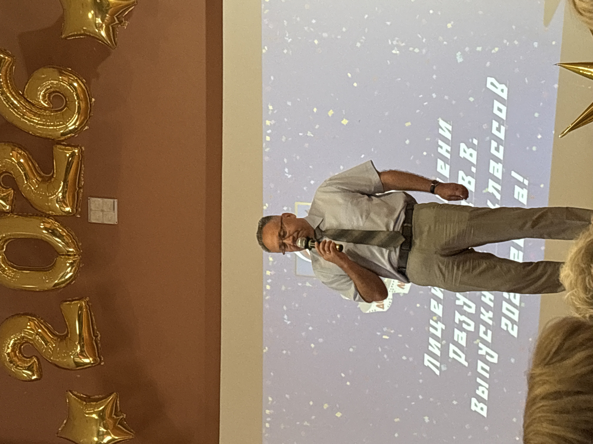 A man in a light blue shirt and tie stands speaking into a microphone in front of a screen. He is wearing glasses and appears to be middle-aged or older, with short, dark hair. The screen behind him displays white text in a vertical orientation, which appears to be in Russian. Some of the visible text includes "В.В. Классов", "Лицей", "РАЗУ", "Выпускн", and "202". The screen is also decorated with confetti. To the left of the man, on a brown wall, are large gold balloon numbers that spell out "202". There are also gold star-shaped balloons. The lighting suggests an indoor event, possibly a graduation or awards ceremony. The man's posture and the microphone indicate he is addressing an audience. The overall atmosphere seems celebratory.