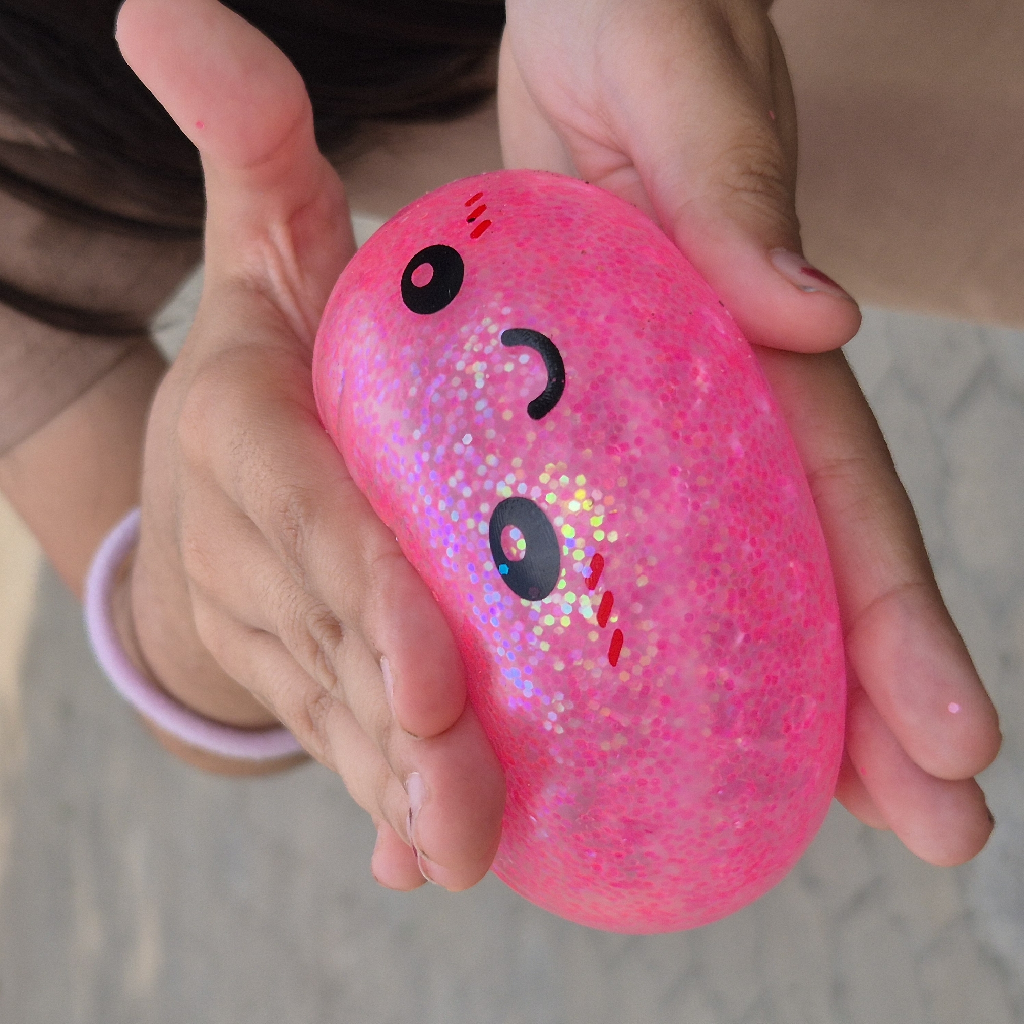 This media file shows a close-up of a person's hands holding a bright pink, glitter-filled stress ball. The ball is oval-shaped and has a cartoon face printed on it, with two black oval eyes and a curved black mouth. There are also three small red dash marks above the face. The hands are light-skinned and appear to be those of a child or young adult, with fingernails painted a light, translucent color. A light pink or purple bracelet is visible on one wrist. The background is out of focus and appears to be an outdoor setting with muted, neutral tones, possibly indicating a neutral or bright day. The lighting suggests it is daytime. The overall emotion conveyed is playful and cheerful due to the bright colors and the cartoonish face on the stress ball. There is no visible text in the media file. The context is Kokshetau, Kazakhstan.