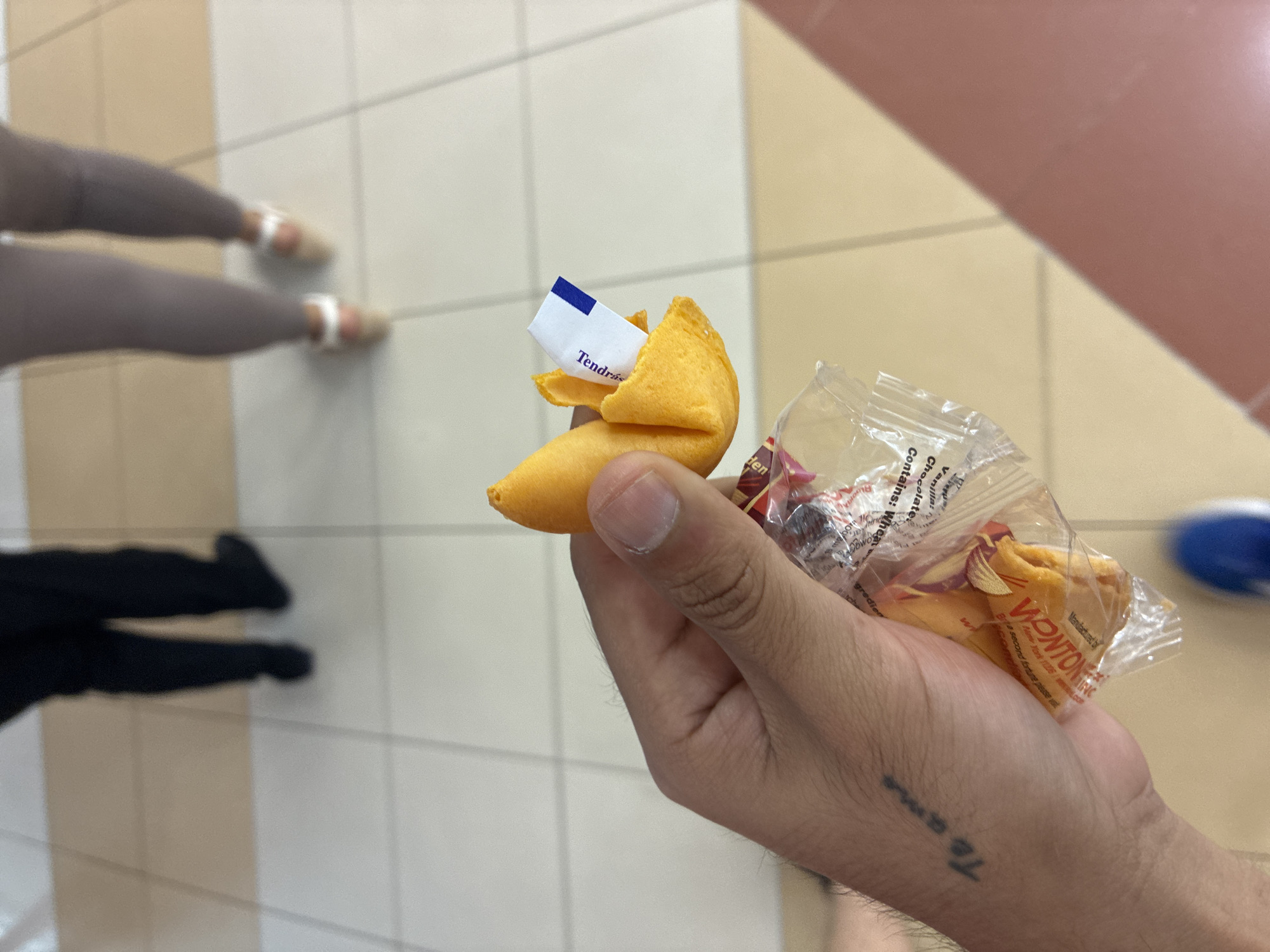 A person holds a fortune cookie in their hand, the wrapper containing another cookie. The fortune sticking out of the cookie reads "Tendrás". The tattoo "Te amo" is visible on the hand. In the background, there are two other people, one wearing black pants and the other wearing grey pants and sandals. The floor is tiled, and the background is blurry. The location cannot be determined from this image, but the fortune is in Spanish, suggesting a Spanish-speaking location. There is nothing in the image that indicates it is in the city of Coatzacoalcos, Mexico.