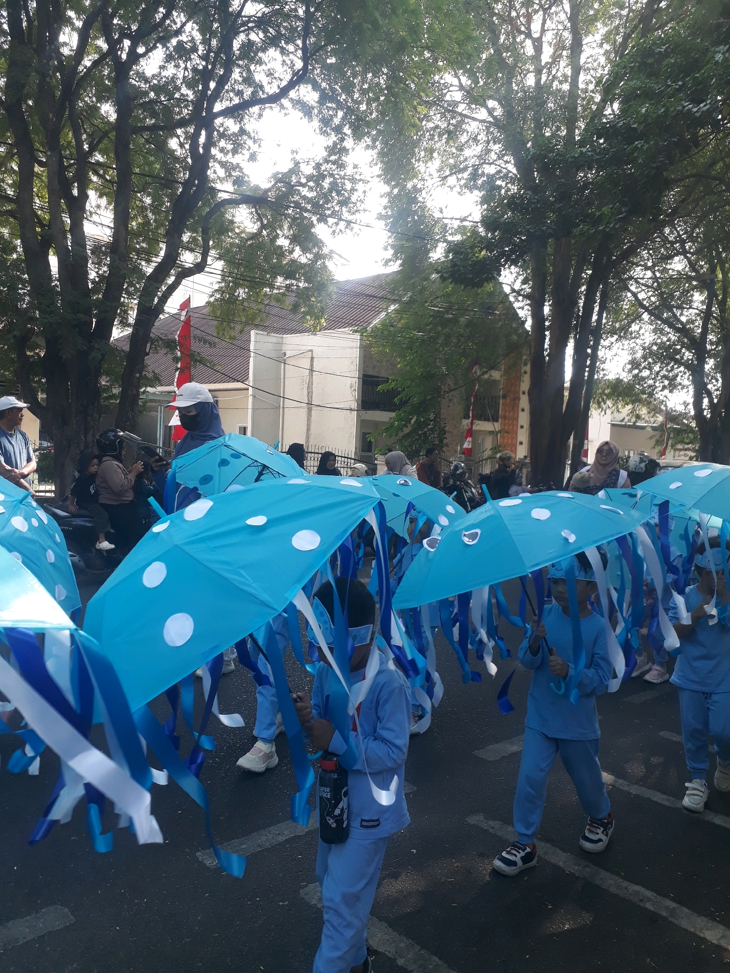 This is a medium shot of a parade in Pangkalpinang, Indonesia. The foreground is dominated by a group of children dressed in light blue uniforms, holding umbrellas that are decorated to resemble jellyfish. The umbrellas are bright blue with white polka dots and have blue and white streamers hanging from them, mimicking the tentacles of a jellyfish. Some of the children are also wearing blue hats or head coverings.
In the background, there are other participants in the parade, some adults and more children, along with spectators. There are trees lining the street, casting shadows on the asphalt. Buildings are visible behind the trees, and a red and white Indonesian flag is displayed on one of them. The atmosphere appears festive, with people gathering to watch the parade. The lighting suggests it is daytime, possibly late morning or afternoon, with sunlight filtering through the trees.