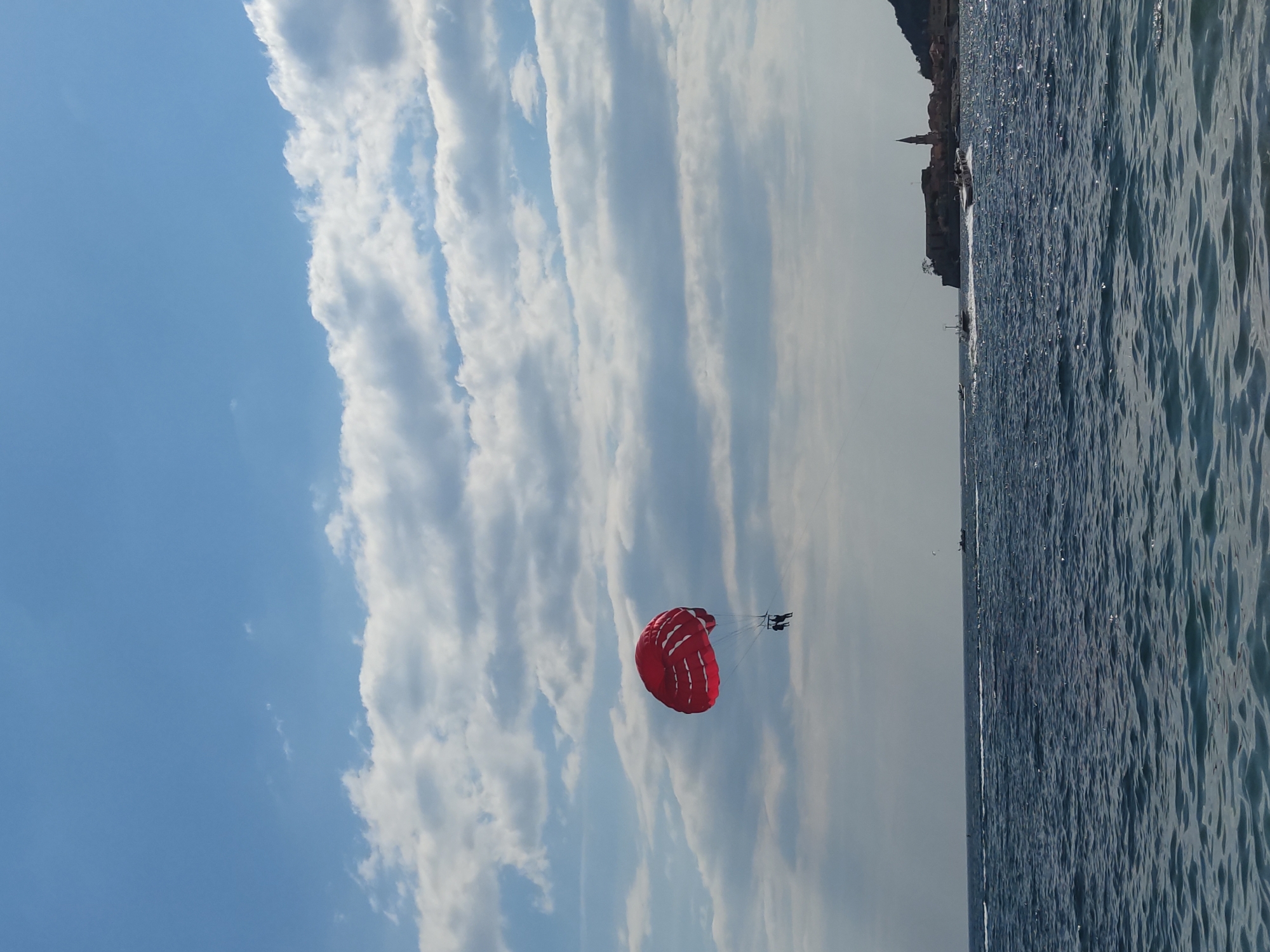 The image shows a red parasail with two people suspended beneath it. The parasail is set against a sky with light blue areas and streaks of white clouds. To the right of the parasail, there is a body of water, possibly a sea or ocean, with visible surface ripples. In the far distance above the water, there is a faint outline of a coastline with a few buildings or structures, including a tower or spire.