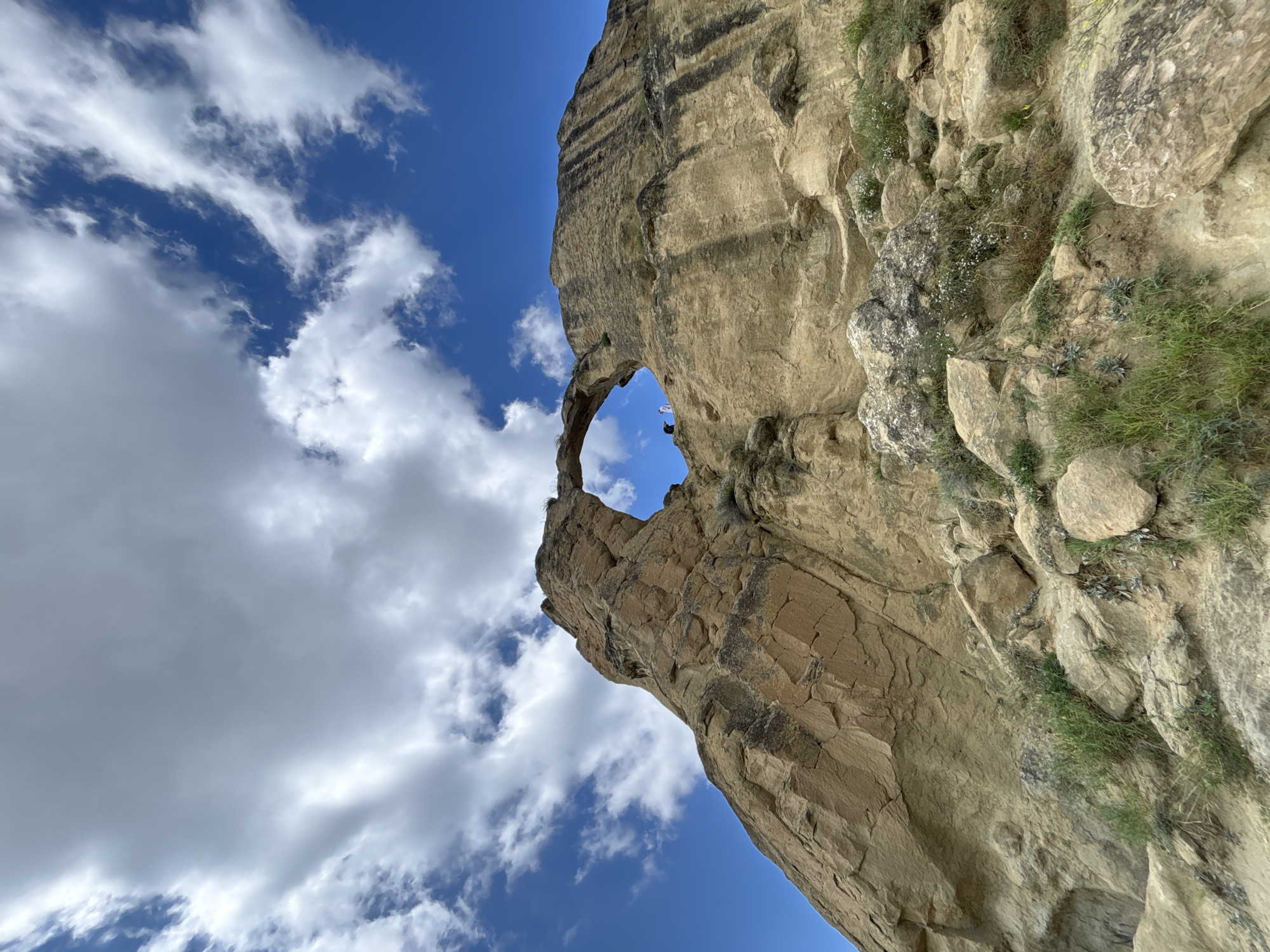 A low-angle shot captures a natural rock arch formation against a bright blue sky dotted with white cumulus clouds. The rock is a light tan color, with visible layers and striations. Sparse green vegetation grows among the rocks at the base of the formation. Two small figures, one wearing white and the other dark clothing, stand near the top of the arch, appearing to be on or near the rock. The arch is roughly circular and overlooks the rocky terrain below. The scene is one of natural geological wonder.