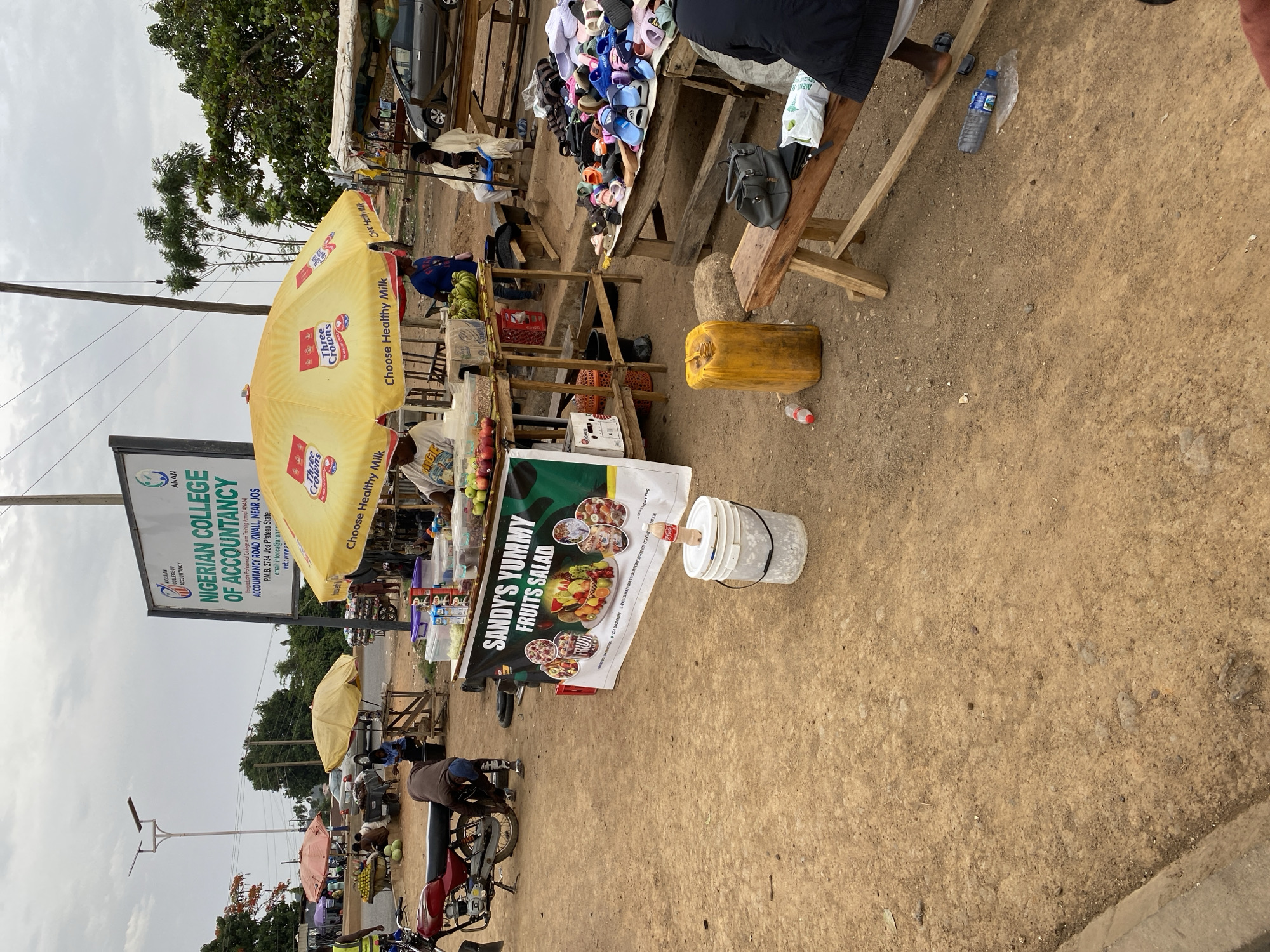 This image captures a vibrant outdoor market scene in Bukuru, Nigeria, under an overcast sky, suggesting daytime. The setting is an unpaved, dusty area, typical of street vending environments.
In the foreground, a prominent yellow umbrella, branded with the "Three Crowns" logo and the slogan "Choose Healthy Milk," shades a stall displaying fresh produce such as apples and bananas. Adjacent to this, a large banner advertises "SANDY'S YUMMY FRUITS SALAD," featuring images of various fruit salad bowls. A white bucket with a red plastic bottle, possibly for beverages, and a yellow jerrycan are also visible on the ground.
To the right, another wooden stall is laden with numerous pairs of colorful slippers and sandals, indicating a footwear vendor. A person is seated behind this stall. Further to the left, another individual is bent over a red motorcycle, seemingly attending to it.
The background features a large white signboard for the "NIGERIAN COLLEGE OF ACCOUNTANCY," which provides specific location details: "ANAN," "Postgraduate Professional College and Training Arm of ANAN," "ACCOUNTANCY ROAD KWALL, NEAR JOS," and "P.M.B. 2734, Jos Plateau State." Additional market activity, including other smaller umbrellas and people, can be seen further down the road, which is lined with trees and utility poles with overhead wires. The scene depicts everyday commerce and life in a bustling, informal market setting.