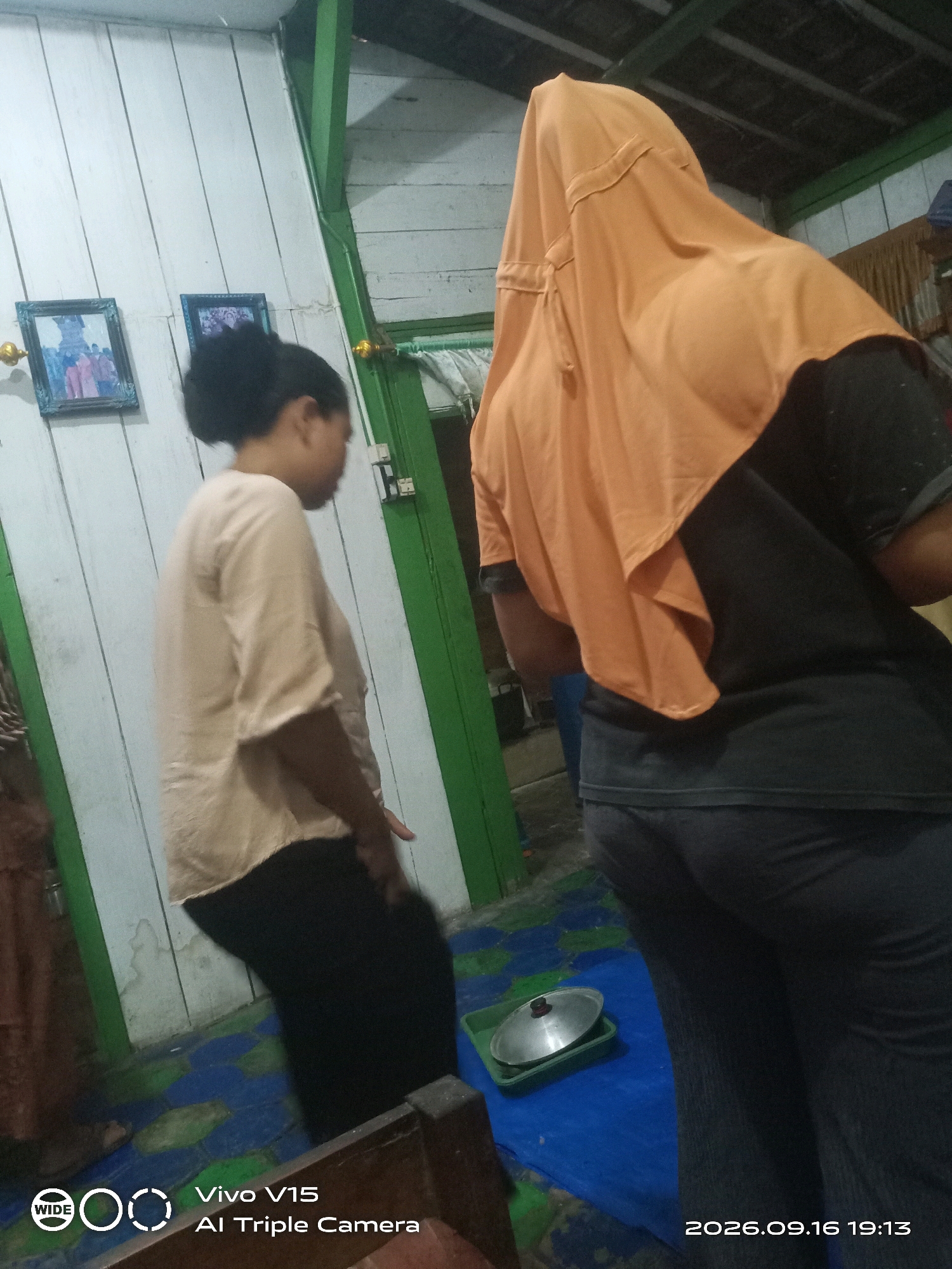 The image captures an indoor scene with two individuals. On the left, a person is visible from the side, wearing a light-colored, loose-fitting shirt and dark pants. Their hair is pulled back. To the right, another person is seen from behind, wearing a dark shirt and dark pants. They are also wearing an orange headscarf that covers their head and shoulders. The background consists of a white wooden wall with two framed pictures hanging on it. A green vertical beam divides the wall. The floor is covered with a blue tarp that has a colorful, abstract pattern. In the foreground, a green rectangular tray with a silver lid is placed on the blue tarp. The timestamp "2026.09.16 19:13" and the text "Vivo V15 AI Triple Camera" are visible in the bottom right and bottom left corners, respectively. The overall impression is that of a casual domestic setting, likely a home in Plosorejo, Indonesia, given the provided context.