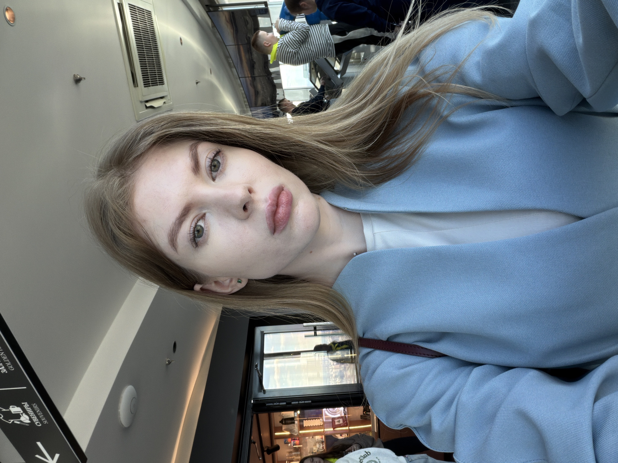 A fair-skinned woman with long, light-brown hair is looking directly at the camera. She is wearing a light blue blazer over a white top. The background suggests an airport or transit hub, with signage, ventilation systems, and seating visible. Through a window behind her, what appears to be a shop is visible.
The location is Khimki, Russia.