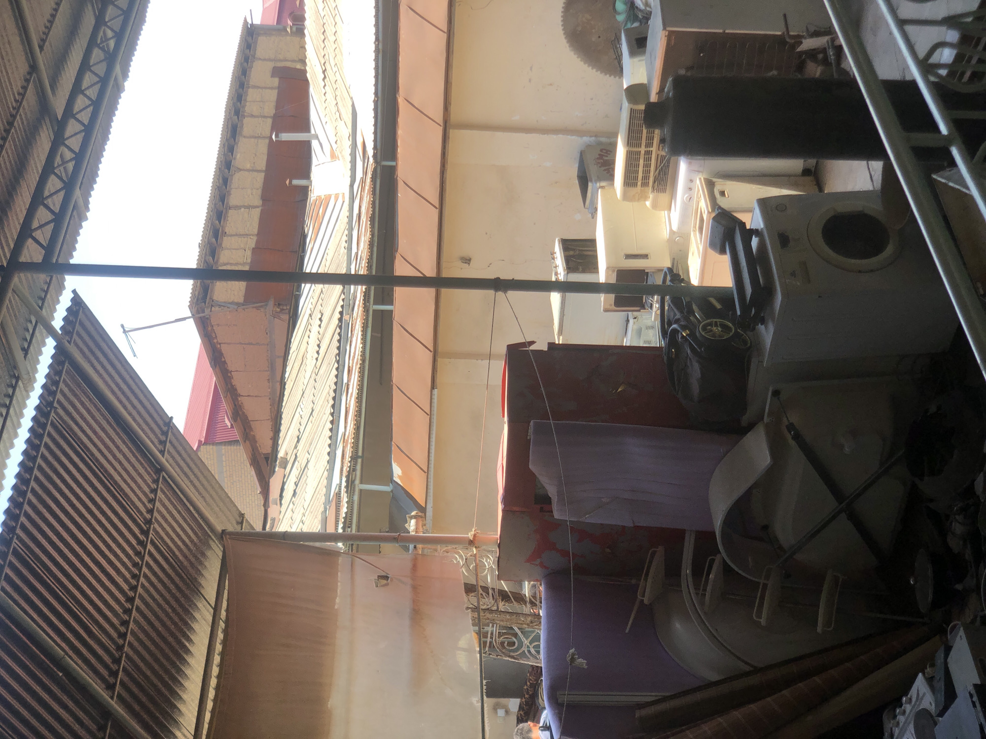 An outdoor storage area in Tashkent, Uzbekistan, is depicted with a variety of discarded appliances and building materials under a corrugated metal roof. The scene is cluttered with items such as washing machines, air conditioning units, large red storage containers, and what appear to be bathtub fixtures. In the background, several buildings with corrugated metal roofs and some with red tile roofs are visible, suggesting an urban or semi-urban industrial or commercial zone. A person is partially visible on the left side of the image, wearing dark clothing and facing away from the camera. The overall impression is of a junkyard or recycling facility.