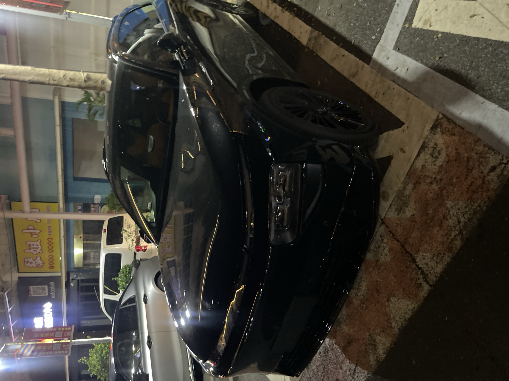 A black car is parked on the side of a street at night, with its headlights off. The car is a modern sedan with a sleek design. The street is paved with asphalt and has white and yellow lines marking parking spaces. There are other cars parked nearby, including a white van and a silver SUV. The background shows a building with illuminated signs, suggesting a commercial area. The scene is set in Panyu District, China. The time of day is night, and the weather appears to be clear. There are no people visible in the image. The overall mood is quiet and still, typical of a street scene at night. The visible text on the signs is in Chinese characters. One sign has yellow lettering on a black background and appears to be a business name or advertisement. Another sign has red lettering on a white background, also likely a business or promotional message. The lighting from the signs and streetlights creates reflections on the car's surface.
