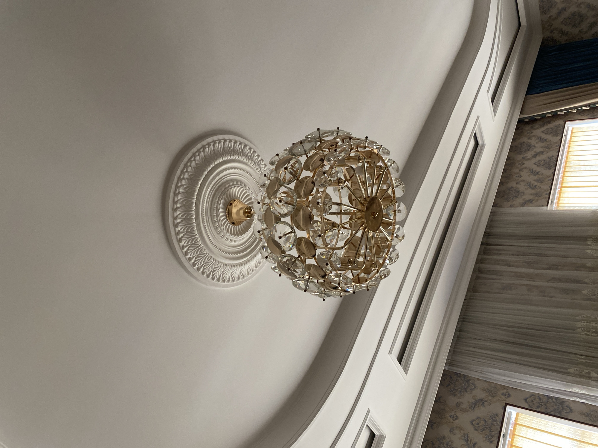 An ornate, crystal chandelier hangs from a white, recessed ceiling with decorative molding. The chandelier is spherical and appears to be made of gold-toned metal and faceted crystals. Below the ceiling, a patterned wallpaper is visible, along with sheer and darker curtains. The scene is indoors, likely in a residential or hotel setting in Turkistan, Kazakhstan, given the context. There are no people or animals present in the image. The lighting suggests it is daytime.