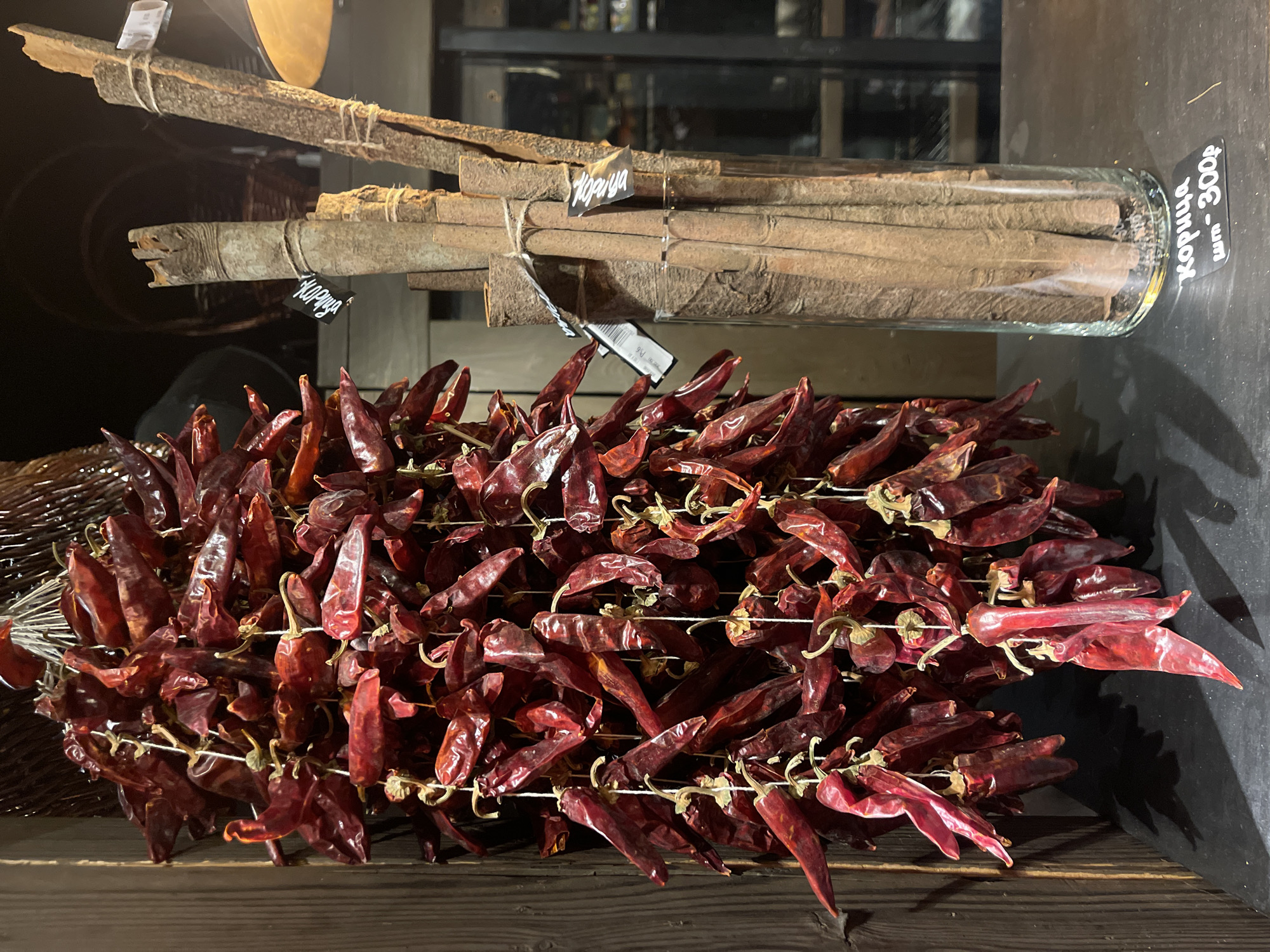 The image depicts a display of culinary items, likely inside a store or market, with a Russian label.
In the foreground, there's a large bunch of dried red chili peppers strung together on a string, creating multiple layers of chili peppers. The chilies appear wrinkled and deep red, typical of dried peppers.
Behind the chilies, on the right side, there is a glass jar filled with cinnamon sticks. A black tag with white writing in Cyrillic is attached to the jar, along with a price label "300 p". The cinnamon sticks appear cylindrical and vary in thickness, with a rough bark texture. Another similar bunch with cinnamon is on the left side.
The scene is set against a dark backdrop with a dimly lit interior visible in the background. The shelf appears to be made of dark wood.