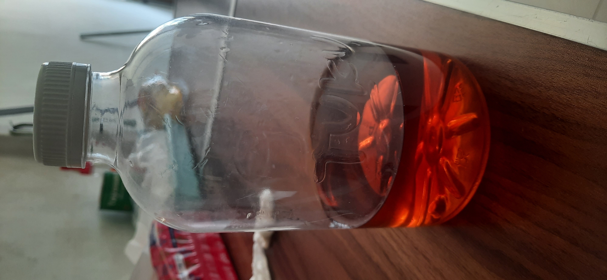 A clear plastic bottle with a gray cap is filled with amber-colored liquid. The bottle is placed on a brown wooden table. The bottle has some text embossed on its side, though it is partially obscured by the liquid and reflections. In the background, a white wall is visible, along with some indistinct objects. The lighting suggests an indoor environment. This scene is located in Cisauk, Indonesia.