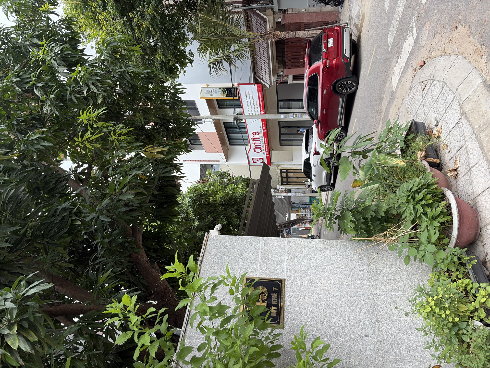 The image depicts a street scene in Đà Nẵng, Vietnam, specifically on Mỹ Khê street. In the foreground, a textured granite wall is visible on the left, bearing a plaque with the inscription "15 Mỹ Khê 7" in gold lettering. Several potted plants, including leafy green varieties and some with dried foliage, are arranged along the sidewalk in front of the wall. A sidewalk made of grey paving stones leads towards a street where two cars are parked. A white SUV is parked closer to the viewer, and behind it, a red SUV is parked further down the street.
Across the street, a multi-story building with a white facade and pink accents dominates the background. A large red banner with white text that reads "ANTIFIRE" and smaller Vietnamese text is prominently displayed on the building's ground floor. Above the banner, a smaller yellow sign with an icon and Vietnamese text is also visible. Trees with dense green foliage overhang the street and the building, providing shade. A palm tree stands to the right of the building. The street appears to be a typical urban street in Đà Nẵng, with paved surfaces and pedestrian walkways. The overall impression is that of a vibrant, lived-in urban environment.