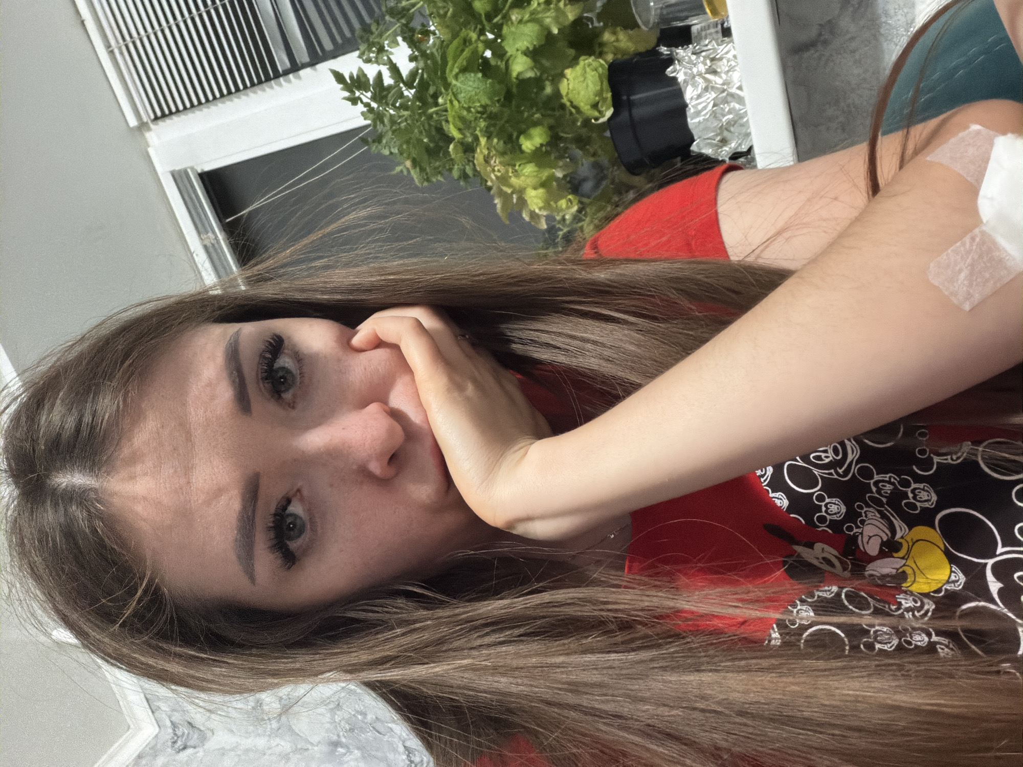 A young woman with long, brown hair and a red t-shirt featuring a Mickey Mouse pattern rests her head on her hand, looking directly at the camera with a somber expression. A bandage is visible on her forearm, held in place by medical tape. The background shows a window with blinds, a plant in a pot, and what appears to be a windowsill. The setting is indoors. No specific location in the city of Япрыково, Russia can be determined from the image.