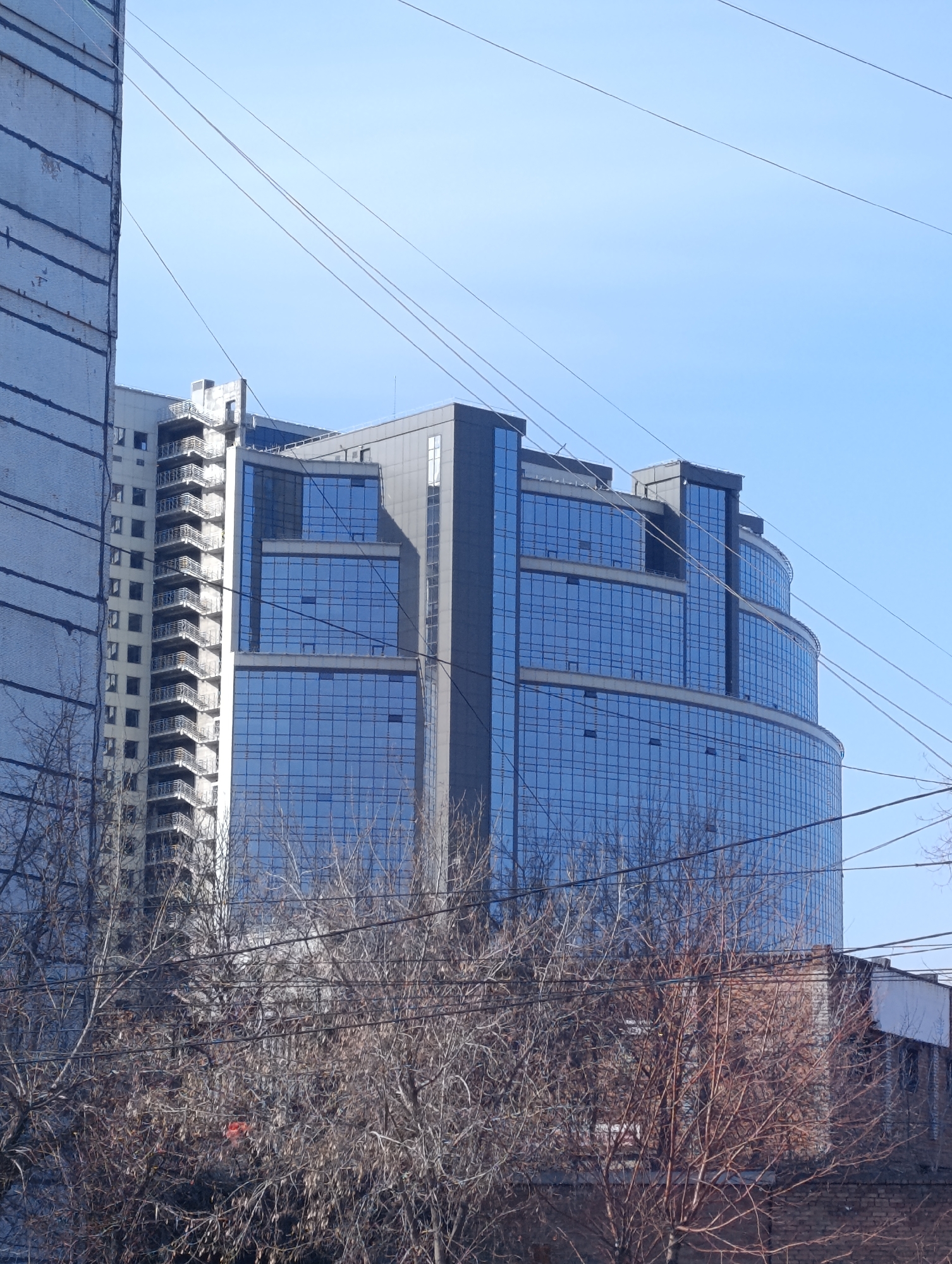 The image shows a modern, multi-story glass building in an urban setting. The building has a curved facade and is divided into sections by horizontal bands. It appears to be located in Krasnoyarsk, Russia. There is another building to the left that looks older and made of brick. Wires, possibly utility lines, run across the sky in the foreground. Bare trees fill the lower part of the image, partially obscuring the buildings. The sky is light blue.
