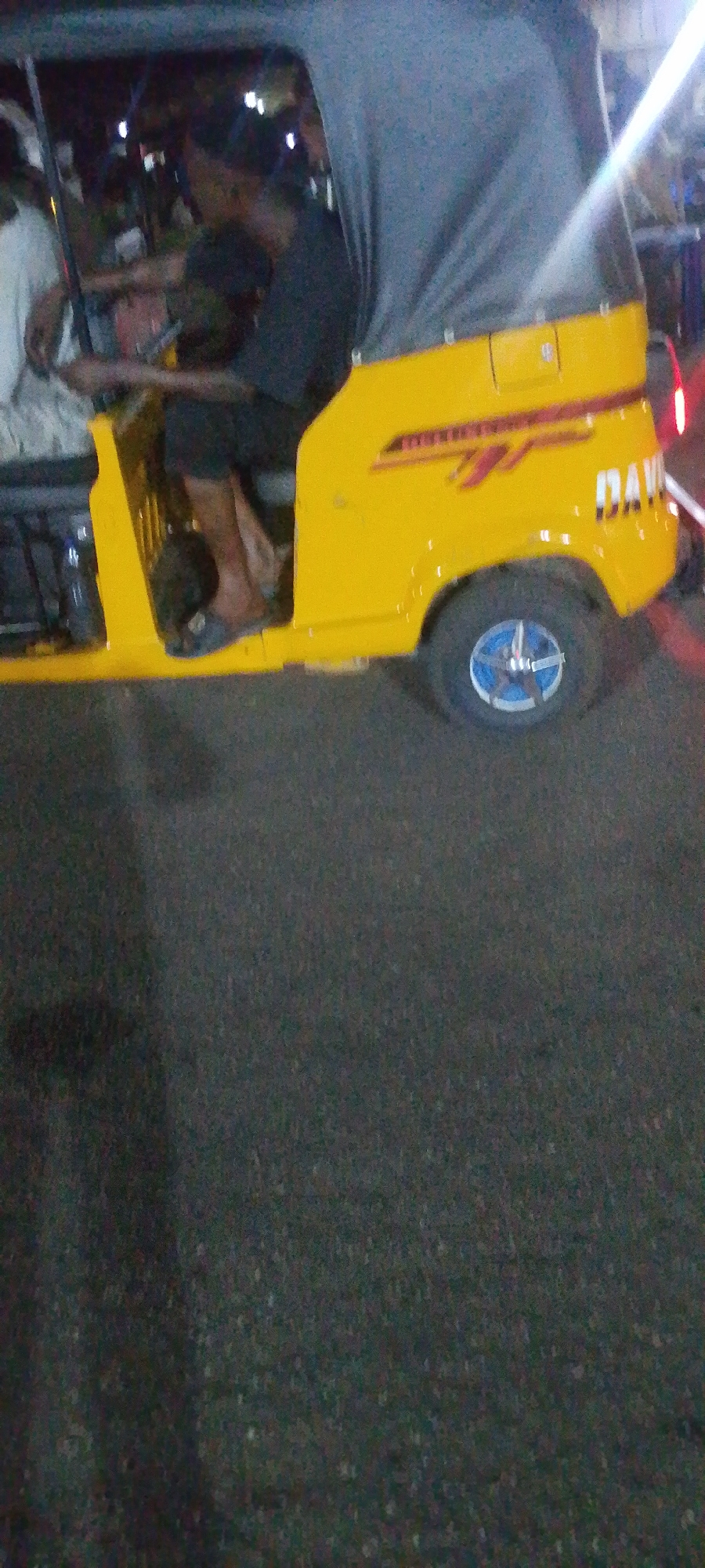 The image depicts a yellow and gray auto rickshaw, commonly known as a "keke" in Nigeria, parked on a dark asphalt surface. The rickshaw features a driver, partially visible, wearing dark clothing and driving barefoot, with a passenger visible to the left. The vehicle's details include blue hubcaps on the rear wheel, a "DAVID" inscription, and red and yellow stripes on its side. The setting appears to be nighttime in an urban environment, potentially the city of Lokoja, Nigeria. There are lights visible in the background, suggestive of street or building illumination. The overall scene suggests a common mode of transportation used in urban Nigerian settings.