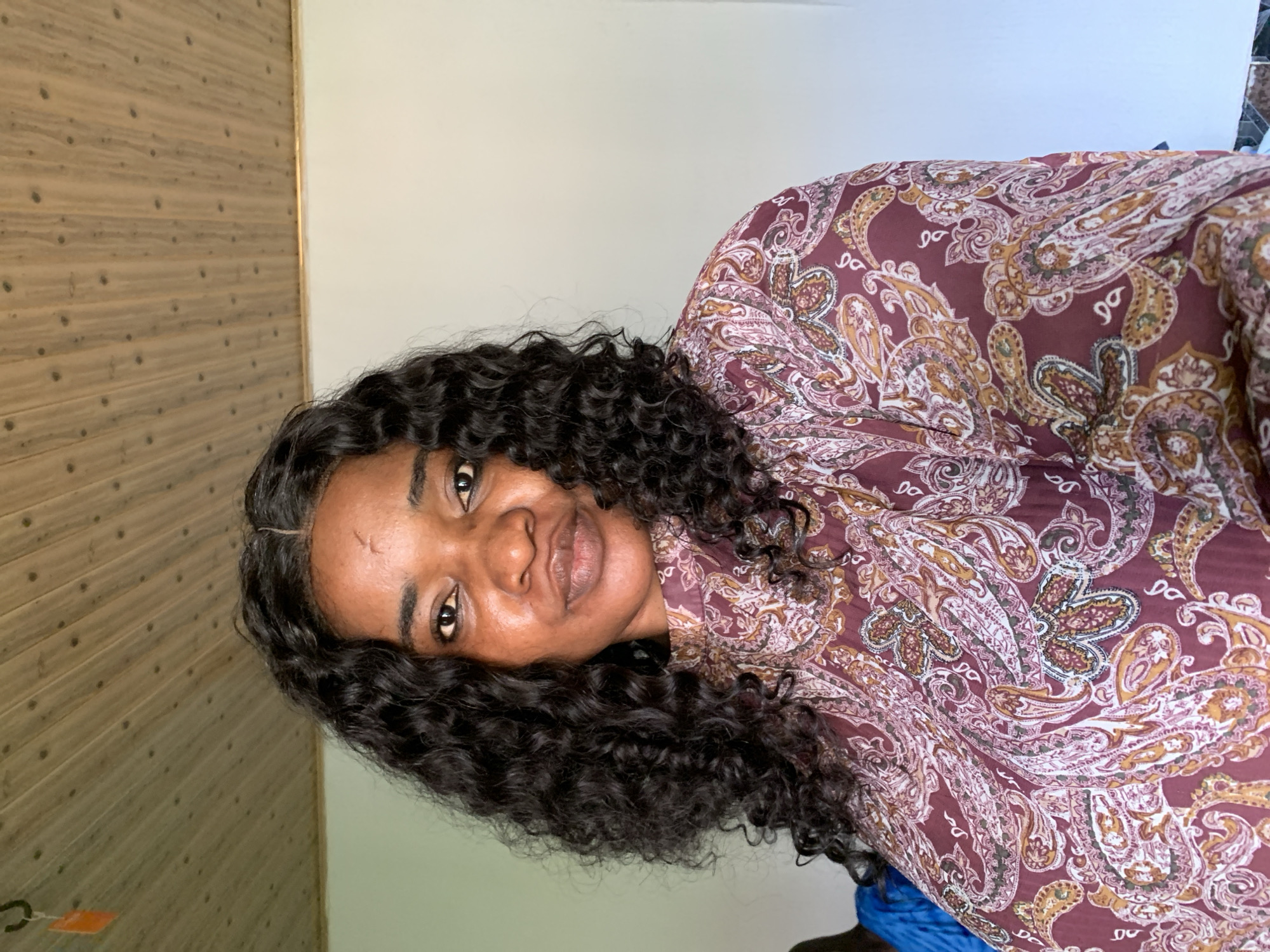 A woman with dark, curly hair and brown skin poses for a portrait indoors. She is wearing a maroon top with a paisley pattern. The background consists of a white wall and a wooden ceiling. The lighting appears to be natural, coming from the front and slightly to the left. There is no indication of the woman's location within Barakin Akawo, Nigeria.