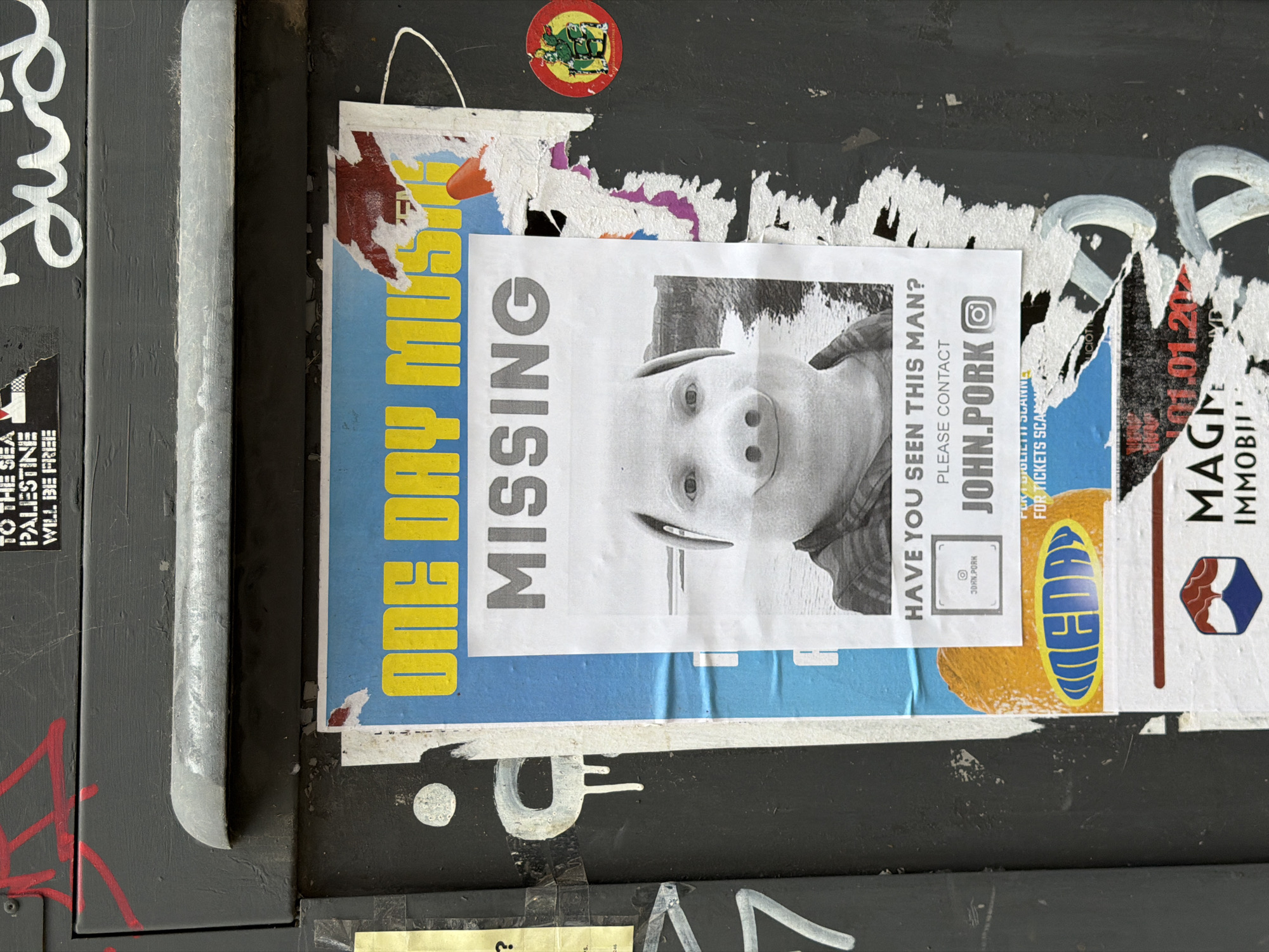 The image shows a section of a wall covered in layers of posters and graffiti. A silver metal bar runs vertically on the left side of the frame. A poster with the words "ONE DAY MUS" in yellow and "MISSING" in gray, with a grayscale image of a person wearing a pig mask is pasted on top of another poster with an orange oval shape and blue letters on it. An adjacent poster asks "HAVE YOU SEEN THIS MAN? PLEASE CONTACT JOHN.PORKO". Graffiti and other fragmented posters are visible above, below, and to the right, with text from other posters showing snippets like "MAGN IMMOBI", and "PALESTINE WILL BE FREE".