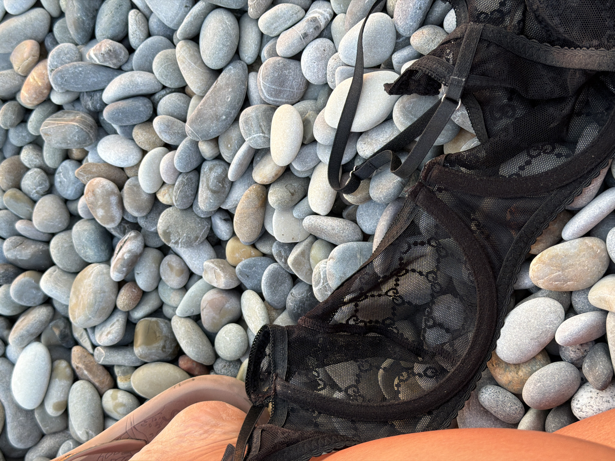 A close-up, high-angle shot shows a black lace bra lying on a bed of smooth, multi-colored pebbles. The pebbles vary in size and shade, ranging from white and light gray to dark gray, blue, and hints of brown and yellow. The bra is partially draped across the pebbles, revealing its delicate lace pattern and straps. To the left of the frame, a portion of a person's tanned leg and hip is visible, with what appears to be a tattoo on the thigh. The scene is set outdoors, likely on a beach in Sochi, Russia, given the abundance of pebbles which are characteristic of the Black Sea coast in that region.