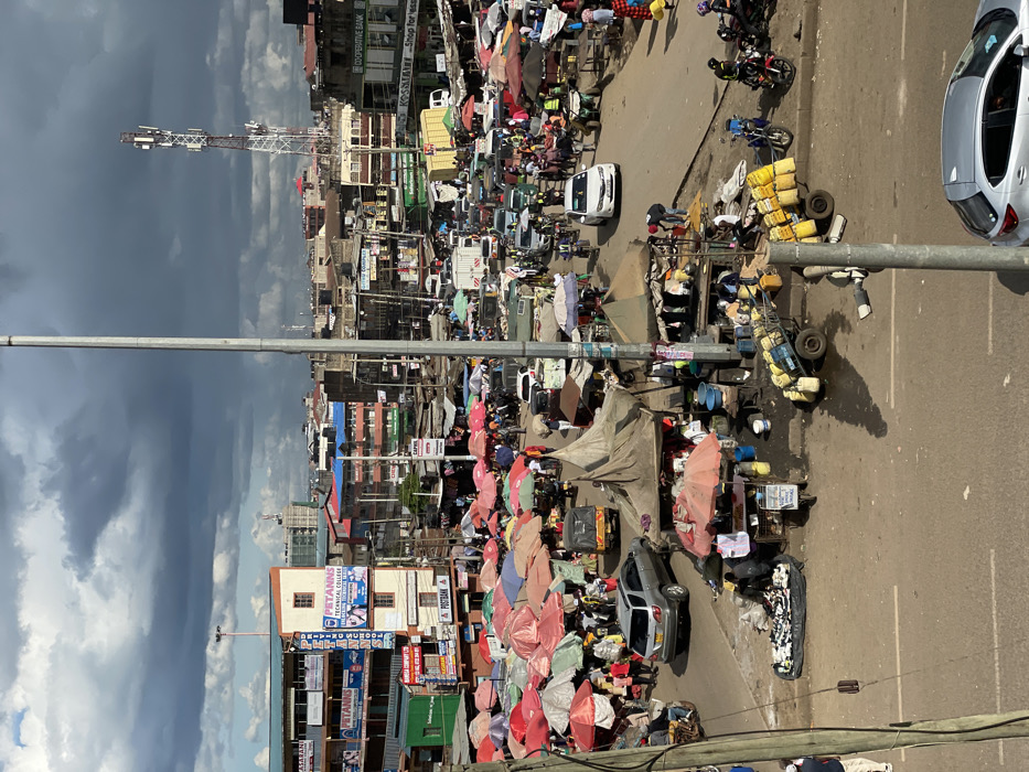 The scene takes place in Githurai, Kenya, at the intersection of two roads, near the coordinates -1.203 latitude and 36.918 longitude. The image shows a bustling outdoor market with numerous vendors selling their wares. The scene is dominated by a variety of stalls, some of which are sheltered by temporary canopies. There are numerous brightly colored umbrellas scattered throughout the market. Hundreds of people are in the market, some are vendors, others are customers, and some are simply walking through. Cars are parked in a haphazard manner on both sides of the road. In the background, there are buildings, including a tall building with the name "PETANNS TECHNICAL COLLEGE" in large lettering. The weather appears partly cloudy with a hint of rain. There is a cell phone tower visible in the background. The street itself is paved and there is a median strip in the middle, separating the two lanes of traffic.