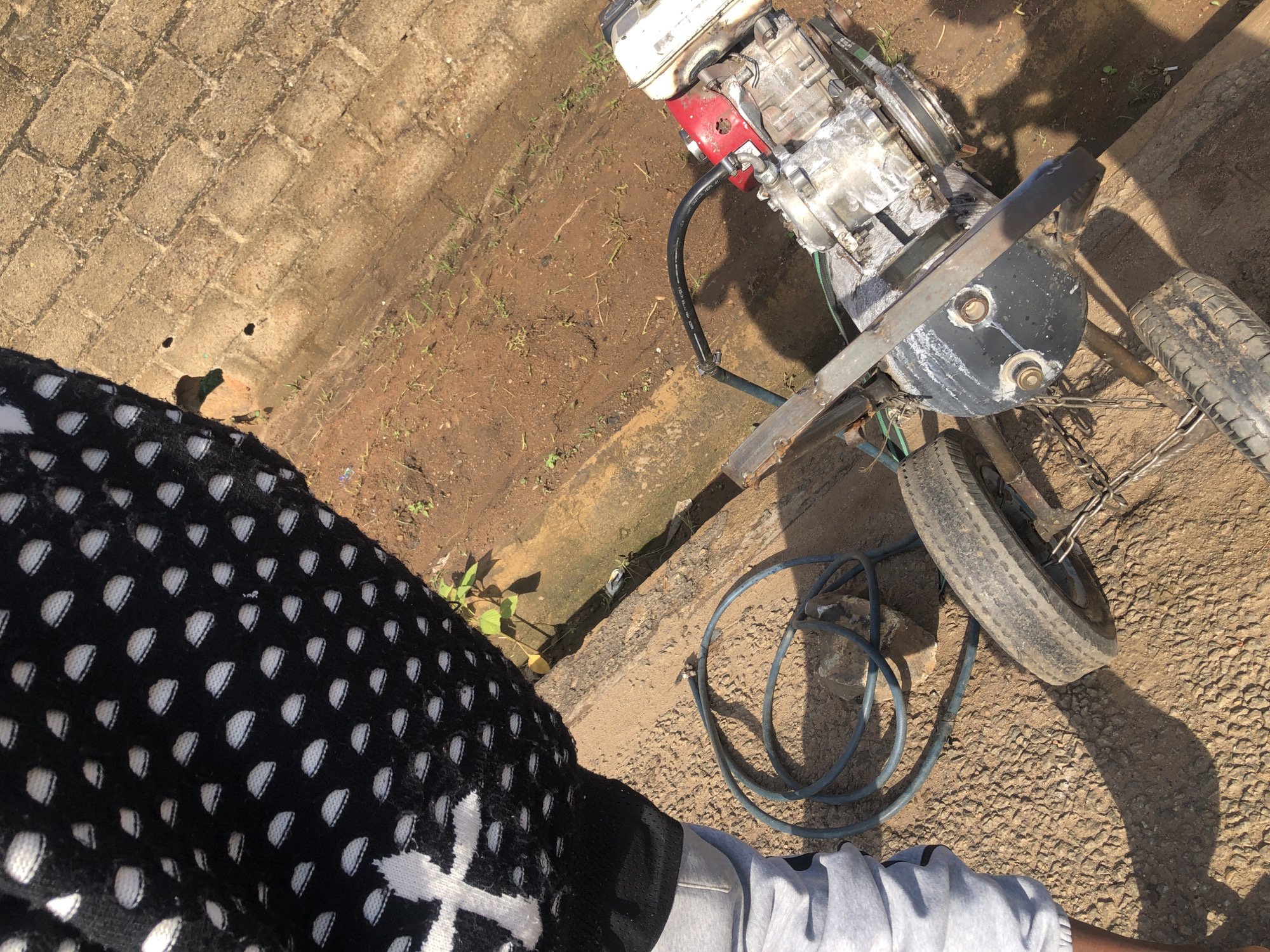 A person wearing a black and white patterned sweater and grey pants stands next to a piece of machinery. The machinery appears to be a portable engine with two wheels, one of which is partially visible. A blue hose is coiled on the ground next to the machinery. The ground is a mix of dirt and gravel, with some loose stones. In the background, there is a brick wall. The scene is outdoors, and the lighting suggests it is daytime. The overall environment appears to be a somewhat unkempt outdoor area, possibly a backyard or a work site.