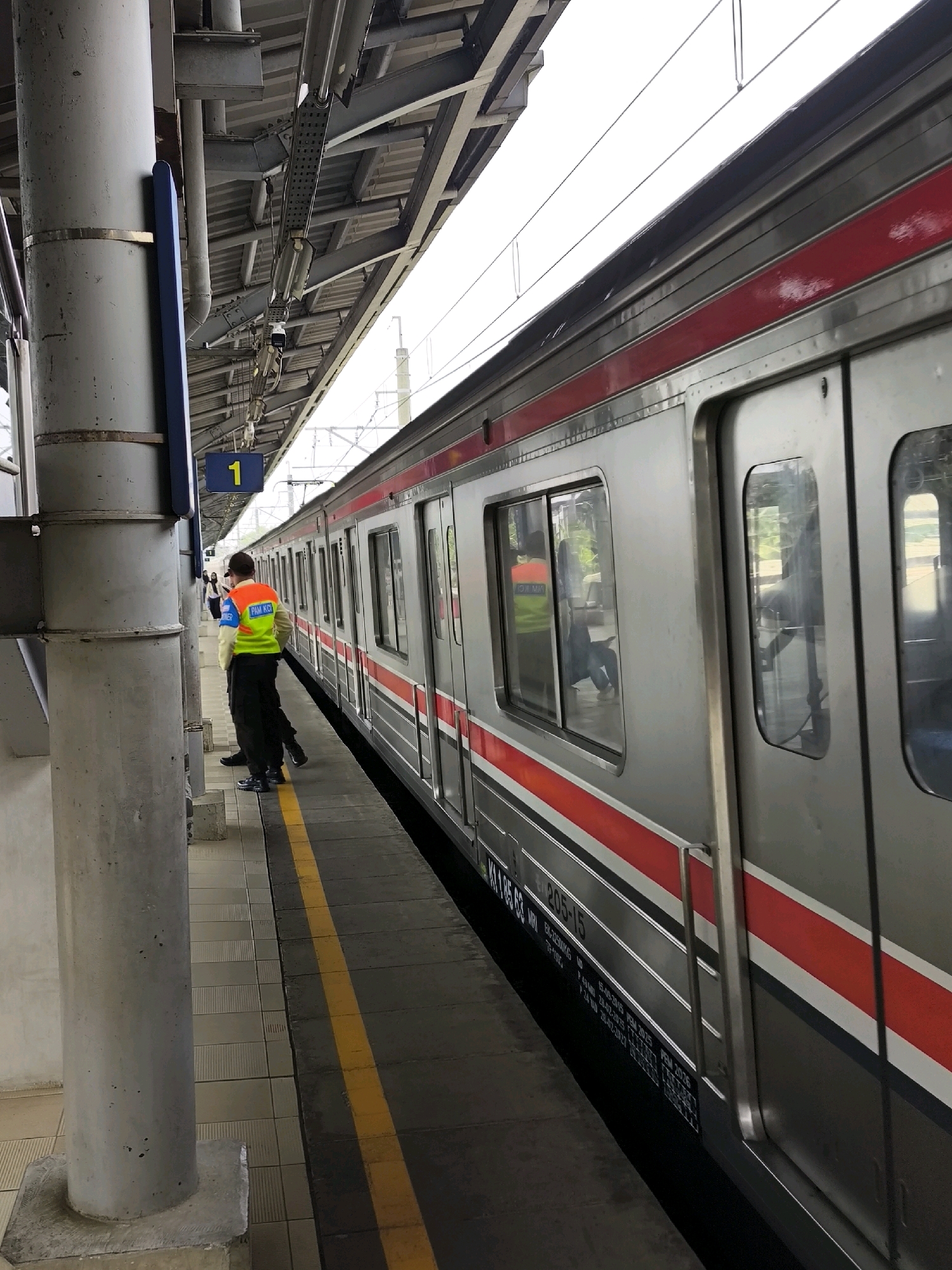 A side view of a commuter train at the Mekarsari, Indonesia train station. The train is silver with a red stripe. The station platform is gray with a yellow line marking the edge. A person wearing a neon orange and green vest stands on the platform, facing the train. The vest has writing on the back that appears to be "PAM KO." Two other figures are visible further down the platform, out of focus. The station has a covered roof supported by metal beams and concrete pillars. A sign with the number "1" in yellow is visible on a blue backing. The sky is overcast.