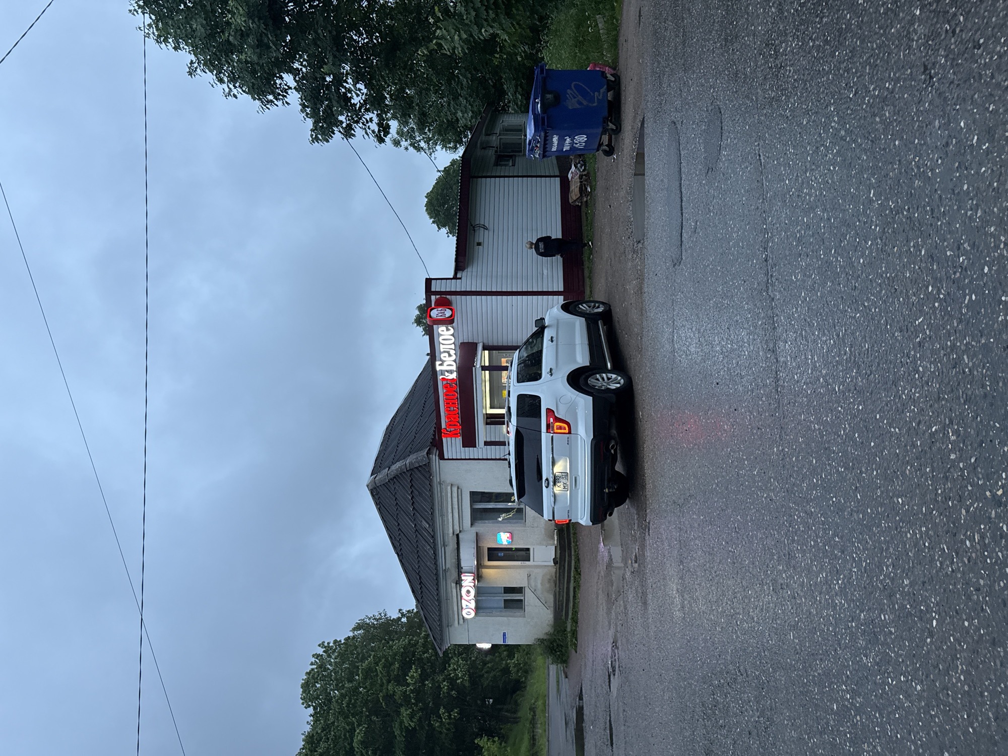 A white Ford Explorer SUV is parked in front of a small, light-colored building with a dark roof, likely a storefront, in Valday, Russia. The building has a sign that reads "Ozon" in neon lights on the left side and another sign with red lettering on the right side that appears to say "Коктейль Белое". A blue dumpster is located to the right of the building, and a person in dark clothing is standing near the rear entrance of the building, facing away from the camera. The ground is wet and appears to be asphalt, with puddles reflecting the dim, overcast sky. Trees are visible in the background on the left and right sides of the image. The overall scene suggests a commercial area on a cloudy day.