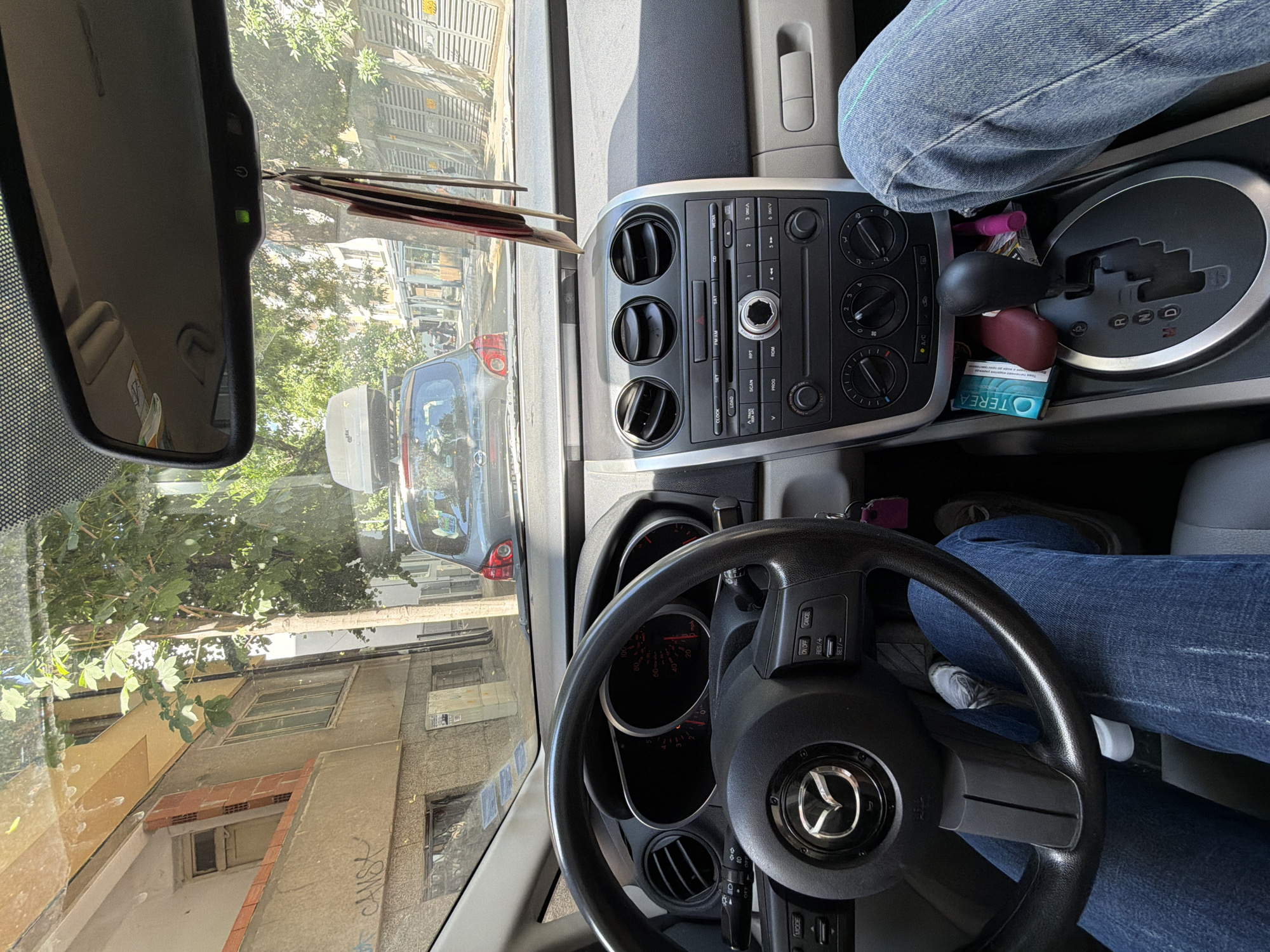 This media file shows the interior of a car, likely a Mazda, viewed from the driver's perspective. The driver's legs, clad in blue jeans, are visible in the foreground, with a hand resting on the steering wheel. The steering wheel itself has Mazda branding. The dashboard features a radio/media system with buttons labeled "MODE", "CLOCK", "LOAD", "SCAN", "RPT", "PROG", "FM AM", "SAT", "CD", and "MEDIA". There are also climate control knobs and buttons with "A/C" visible. A gear shift lever is present, indicating an automatic transmission, with markings for "P", "R", "N", and "D". A pack of "OTEREA" cigarettes is visible near the gear shift.
Outside the car, through the windshield, a street scene is depicted. Buildings with windows and balconies line the street, with trees providing some shade. The architecture suggests an urban environment, and the presence of graffiti on one of the buildings ("CANSK") adds to this context. The lighting indicates it is daytime, likely sunny, given the shadows and brightness. The overall scene suggests the car is either parked or stopped in traffic on a street in Plovdiv, Bulgaria. The image is oriented vertically, with the top of the car's interior appearing at the bottom of the frame.