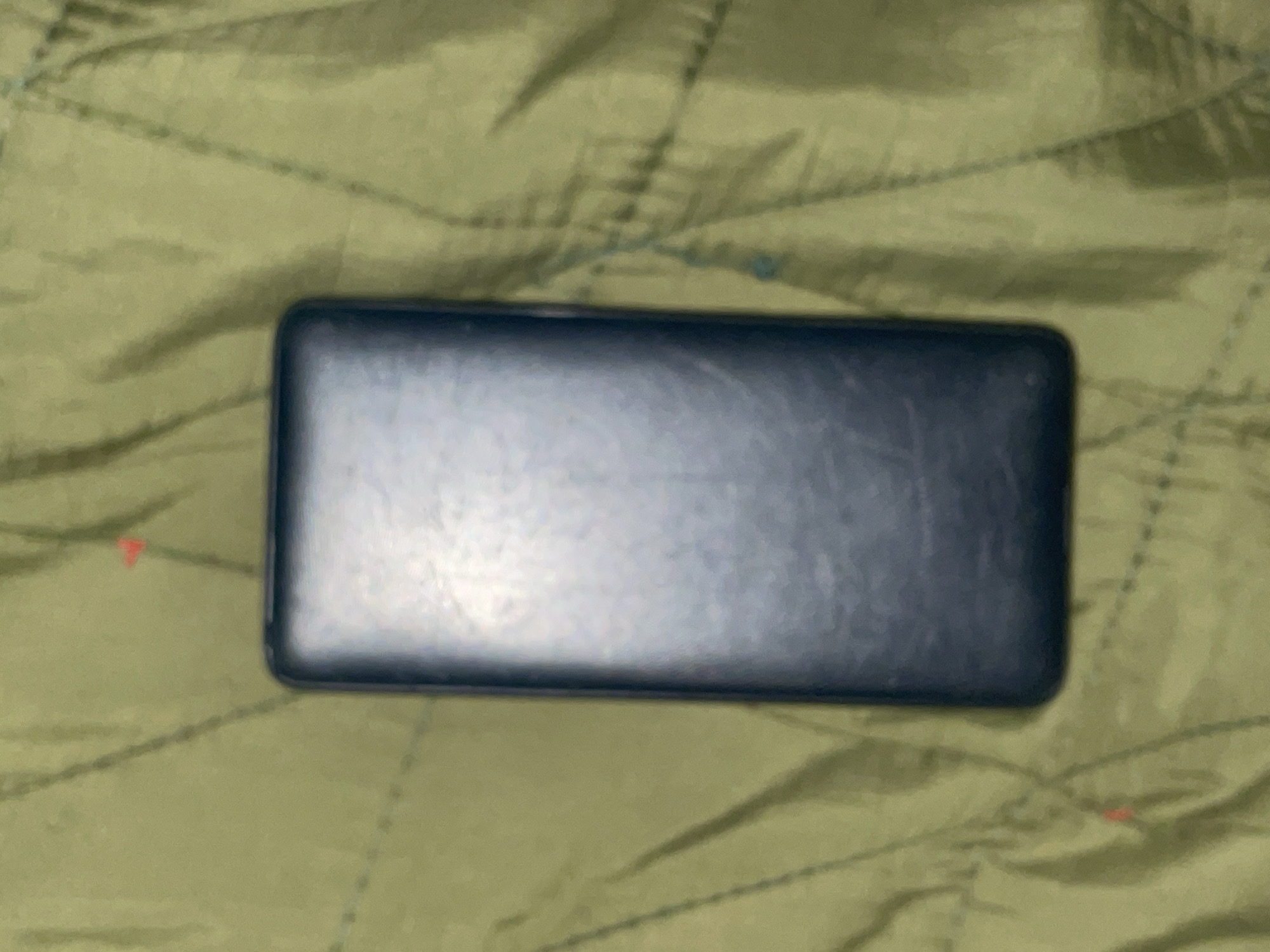 This is a medium-shot, overhead photograph of a dark, rectangular object resting on a textured, olive-green surface. The object appears to be a phone or a similar electronic device, lying horizontally in the center of the frame. Its surface is smooth and shows some subtle reflections, suggesting it might be made of glass or plastic. The green background has a quilted or padded texture with a pattern of diagonal lines and occasional darker shadows. There are a few small, indistinct red and blue marks scattered on the green surface, possibly minor blemishes or color variations. The lighting in the image is somewhat diffused, creating soft shadows and a slightly muted color palette. The overall scene is static, with no apparent activity or human presence. The context of Jalingo, Nigeria, is provided, but there are no specific visual cues within the image to confirm this location. The time of day and weather are not discernible. No text is visible.
