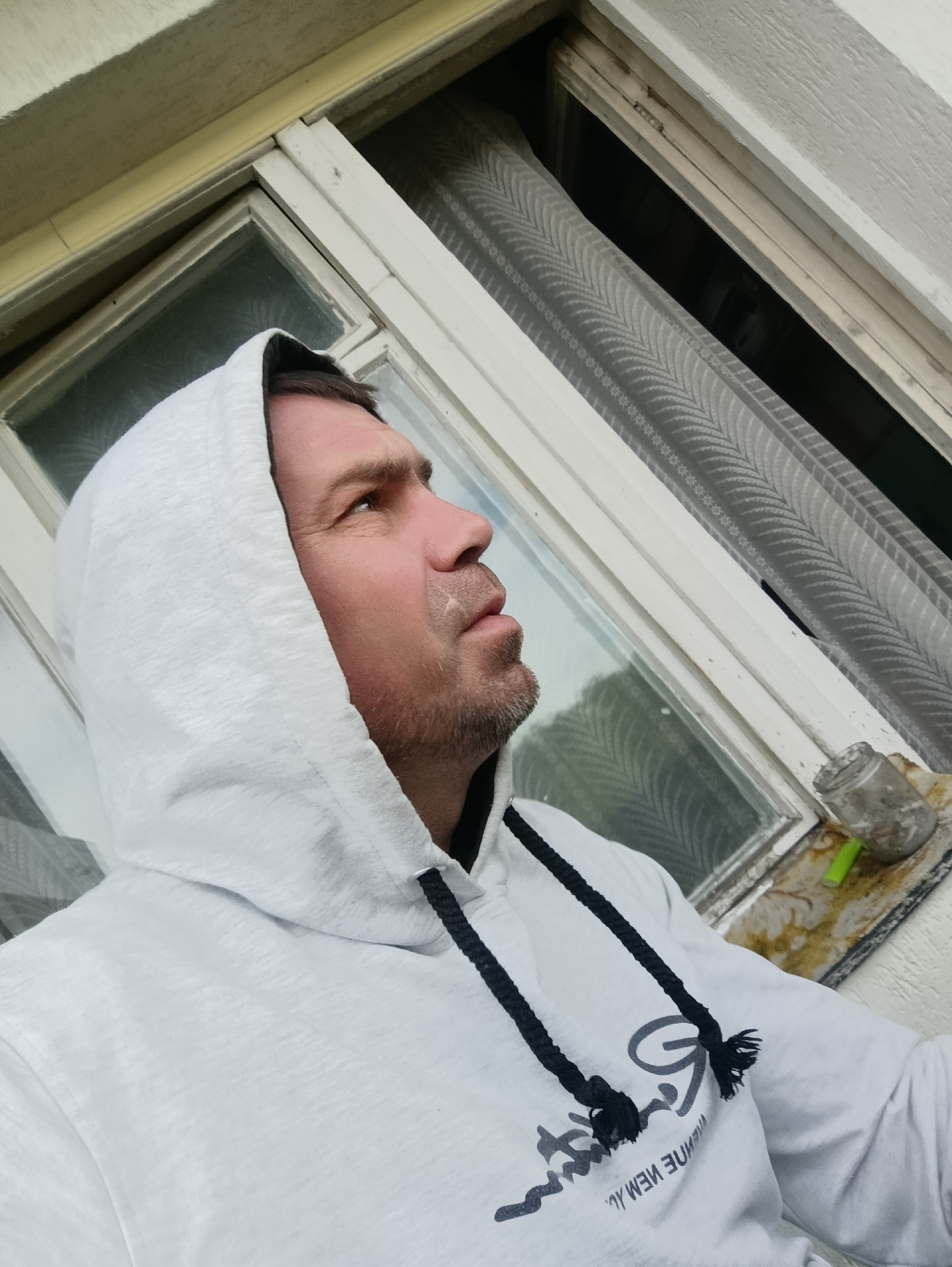 A medium shot shows a man wearing a white hooded sweatshirt with black drawstrings. He is looking up and to the right, with his mouth slightly open. He has short, brown hair and stubble. There is a light-colored mark on his upper lip, possibly paint or dust. The man is positioned in front of a white-framed window with a patterned gray curtain behind it. The window appears to be part of an exterior wall. A small, dirty glass jar with a green object inside sits on the windowsill to the right. The overall scene suggests an outdoor or semi-outdoor environment, potentially a building exterior in Ladyzhyn, Ukraine.