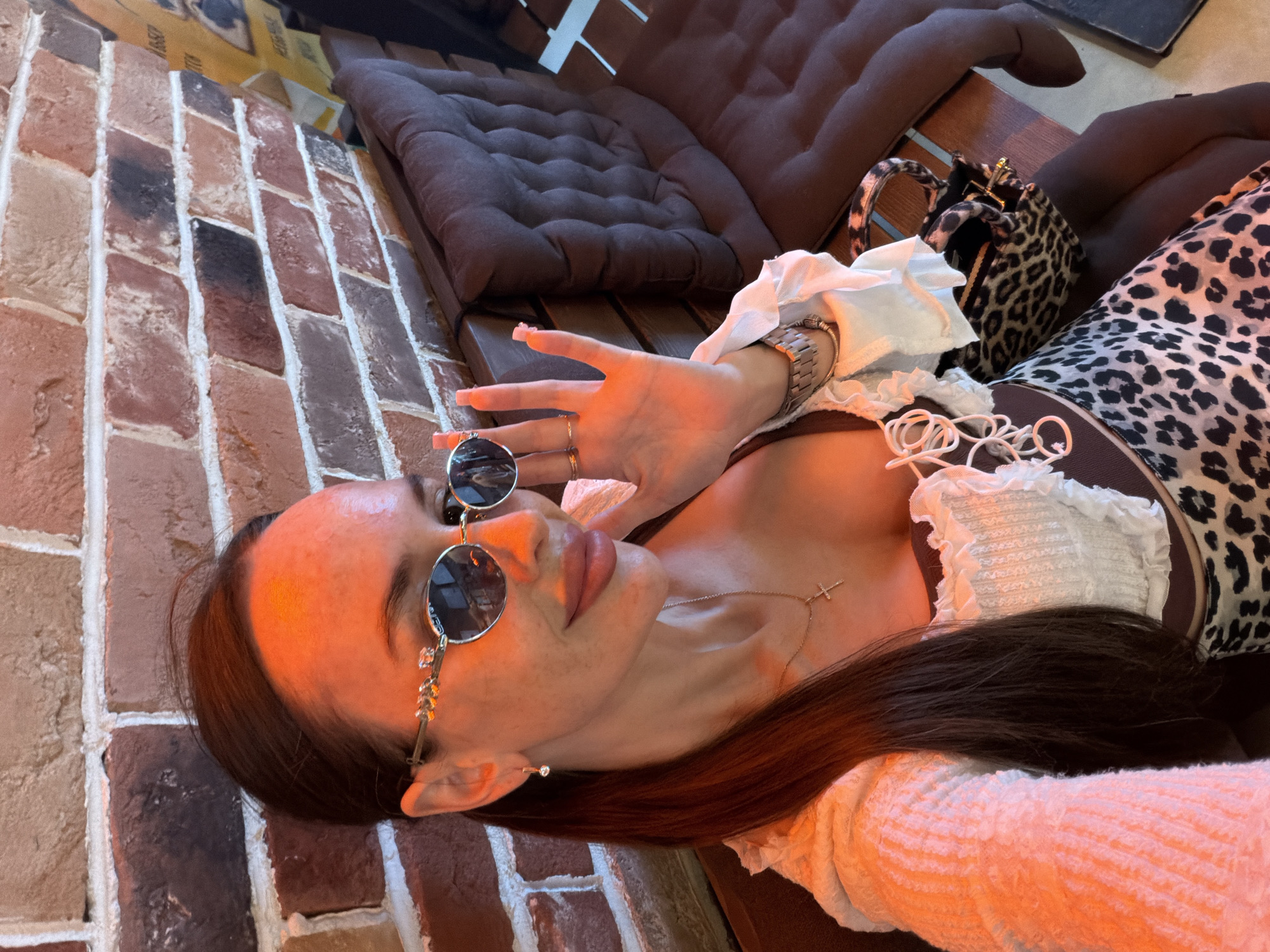 The image depicts a single female individual inside an establishment, likely a cafe or restaurant, located in the city of Saint Petersburg, Russia. The individual, appearing to be in her 20s or 30s, is seated on a dark brown upholstered bench with button tufting. She has long, dark brown hair pulled into a ponytail and is wearing oval-shaped sunglasses with decorative, possibly bejeweled, frames. Her face shows visible freckles, and her lips are full with a light-colored lipstick. She wears a gold chain necklace with a small cross pendant and a small, clear stone earring in her left ear. Her attire consists of a white, ruffled camisole or top, layered under a light-colored, possibly white or light pink, long-sleeved textured knit garment. A leopard print skirt or dress is visible below her waist. On her left wrist, she wears a metallic watch, and multiple rings adorn her right hand, which is raised with the palm facing forward, fingers extended. Her fingernails are long, rectangular, and painted a light, neutral color. Adjacent to her on the bench is a small leopard print handbag with dark handles. The background features an interior brick wall with varied red, brown, and dark-colored bricks and white mortar. A dark wooden surface or armrest runs along the base of the brick wall, in front of the seating. Warm, artificial lighting casts an orange hue across her face and the immediate surroundings. A blurry yellow object, possibly a poster, is partially visible in the upper right background.
