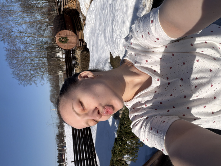 A young adult female is depicted in a selfie, taken outdoors in Shipulino, Russia, under a clear blue sky. She presents with closed eyes, a kiss-like facial expression with pursed lips, and her hair pulled back. She wears a white short-sleeved top adorned with small red heart patterns and a thin gold-colored necklace and matching link earrings. Sunlight casts shadows, illuminating her right side. The background features a snow-covered landscape indicative of winter or early spring. A dark wooden fence traverses the mid-ground. To the left, a snow-dusted field extends towards distant houses and bare deciduous trees. To the right, a prominent cylindrical wooden barrel sauna with a door featuring a green wreath and a metal stovepipe is visible, resting on a wooden platform. A stack of cut firewood is situated behind the sauna. The ground is largely covered in snow, with patches of exposed earth and dormant vegetation.