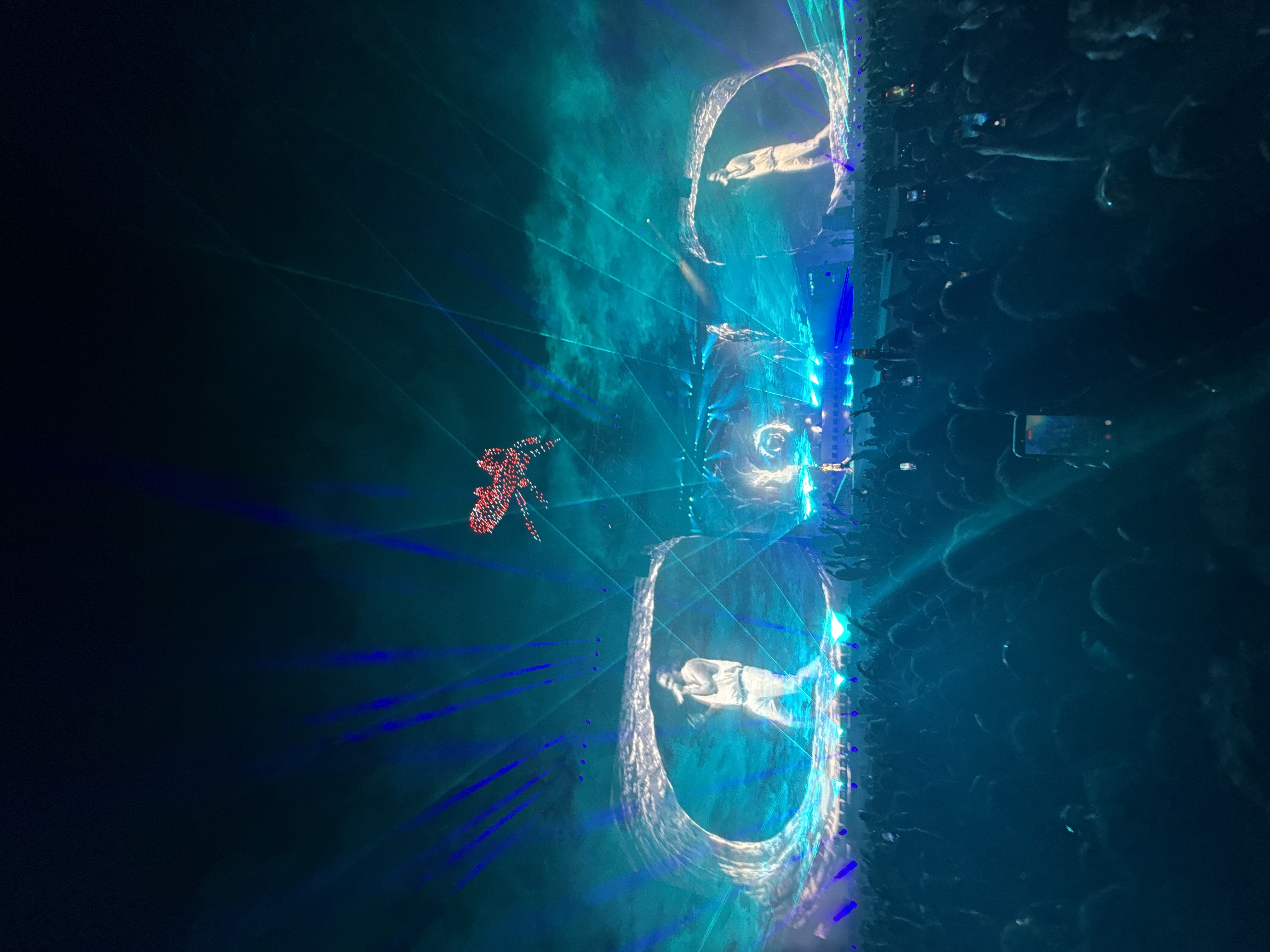 The image shows a concert in Indio, United States, featuring a large crowd of people. The stage is elaborate with several screens displaying performers. Above the stage, there is a formation of what appear to be illuminated drones forming a distinct shape. The stage lighting and effects create a vibrant, energetic atmosphere.