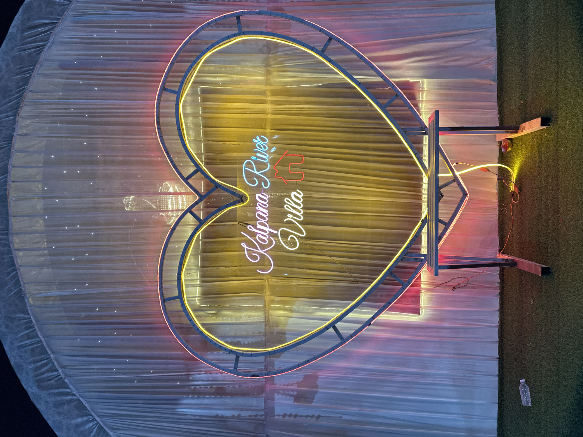 The image shows a brightly lit heart-shaped sign with the words "Kalpana River Villa" in neon lettering. The sign is constructed of metal tubing and illuminated with yellow and red neon lights, and it is placed in front of a translucent white curtain backdrop. The setting appears to be outdoors on a grassy area, possibly in the city of Shahapur, India. A water bottle is visible on the grass to the lower right of the scene.