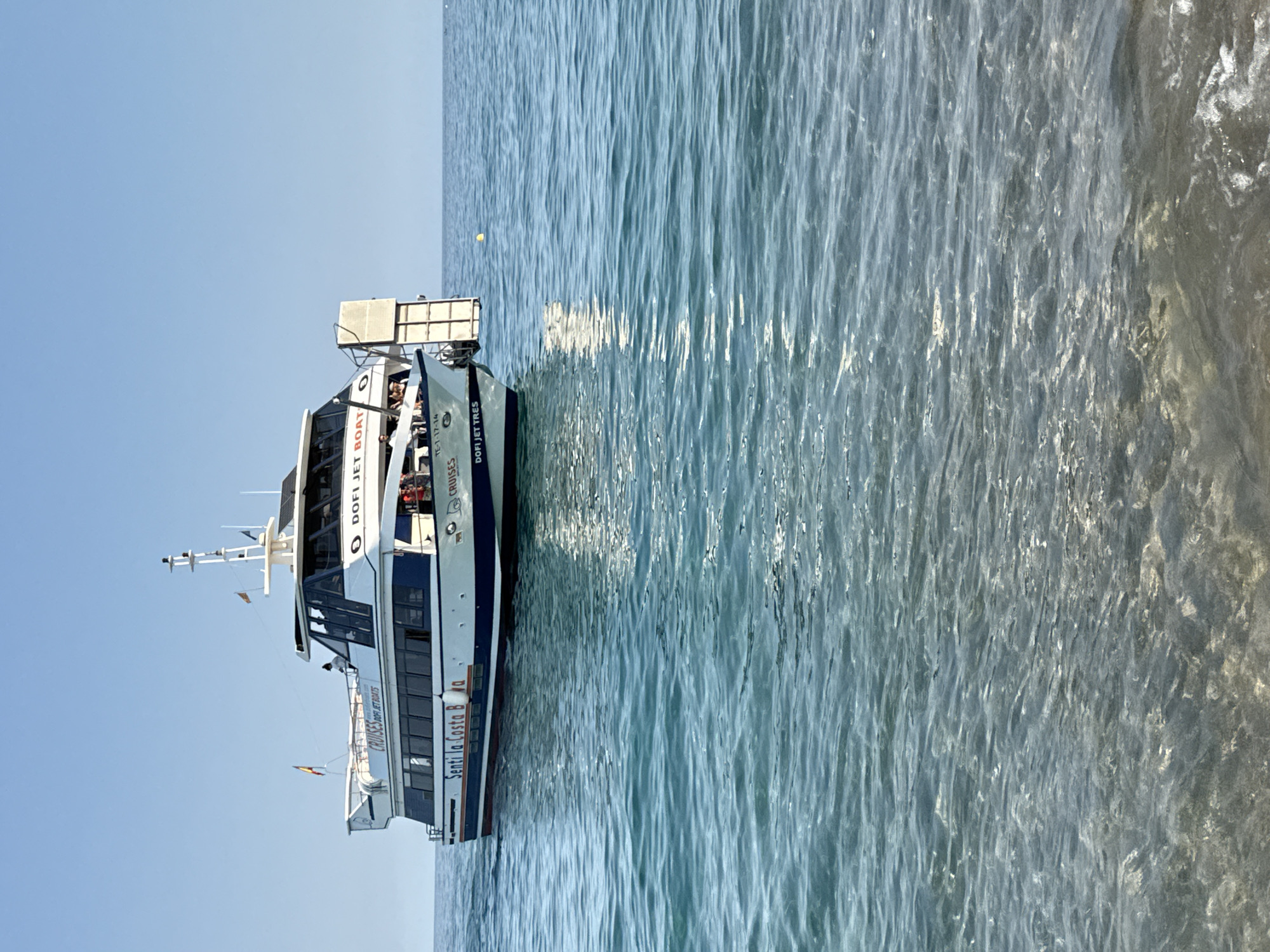 A modern, white and blue passenger boat, named "Dofi Jet Boat" and "Dofi Jet Tres", is cruising on the clear blue Mediterranean Sea off the coast of Blanes, Spain. The boat has multiple decks and windows, with some passengers visible on the upper deck. The water is calm with gentle waves, and the sky is clear and blue. The shoreline is not visible, but the boat is close enough to shore that the seabed is visible through the clear water in the foreground.