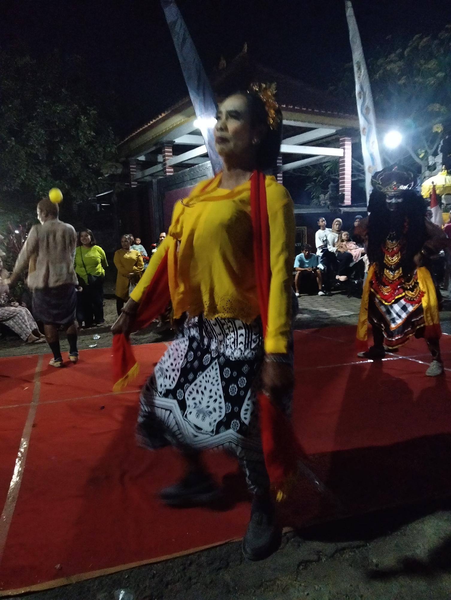In Sumbersewu, Indonesia, a cultural performance unfolds at night on a red carpeted stage. A woman, adorned in a bright yellow top with a red sash, performs traditional dance, her patterned skirt swirling. In the background, another performer, dressed in a dark, elaborate costume with facial mask, participates in the event. A crowd of onlookers, seated and standing, observes the spectacle. A man with a yellow ball balanced on his head is also visible. The scene is illuminated by artificial lights.