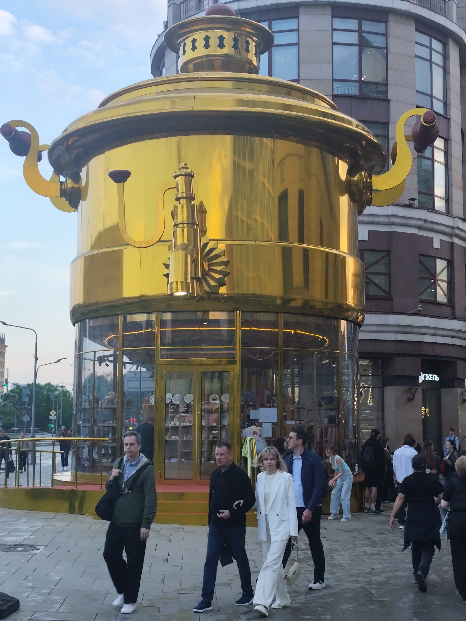 This is a daytime outdoor shot featuring a large, golden samovar-shaped building, standing prominently on a pedestrian area in Moscow, Russia. The building has a glass-enclosed base where a shop likely sells samovars and other goods. The golden samovar structure has handles and spouts.
A group of people is walking around the building, including a man in a green jacket, a man in a black jacket, and a woman in a white suit. Other pedestrians surround them.
In the background, there's a multi-story building, and across the street, a lamppost and traffic lights. The sidewalk is paved. The overall environment is urban and busy with pedestrian activity.