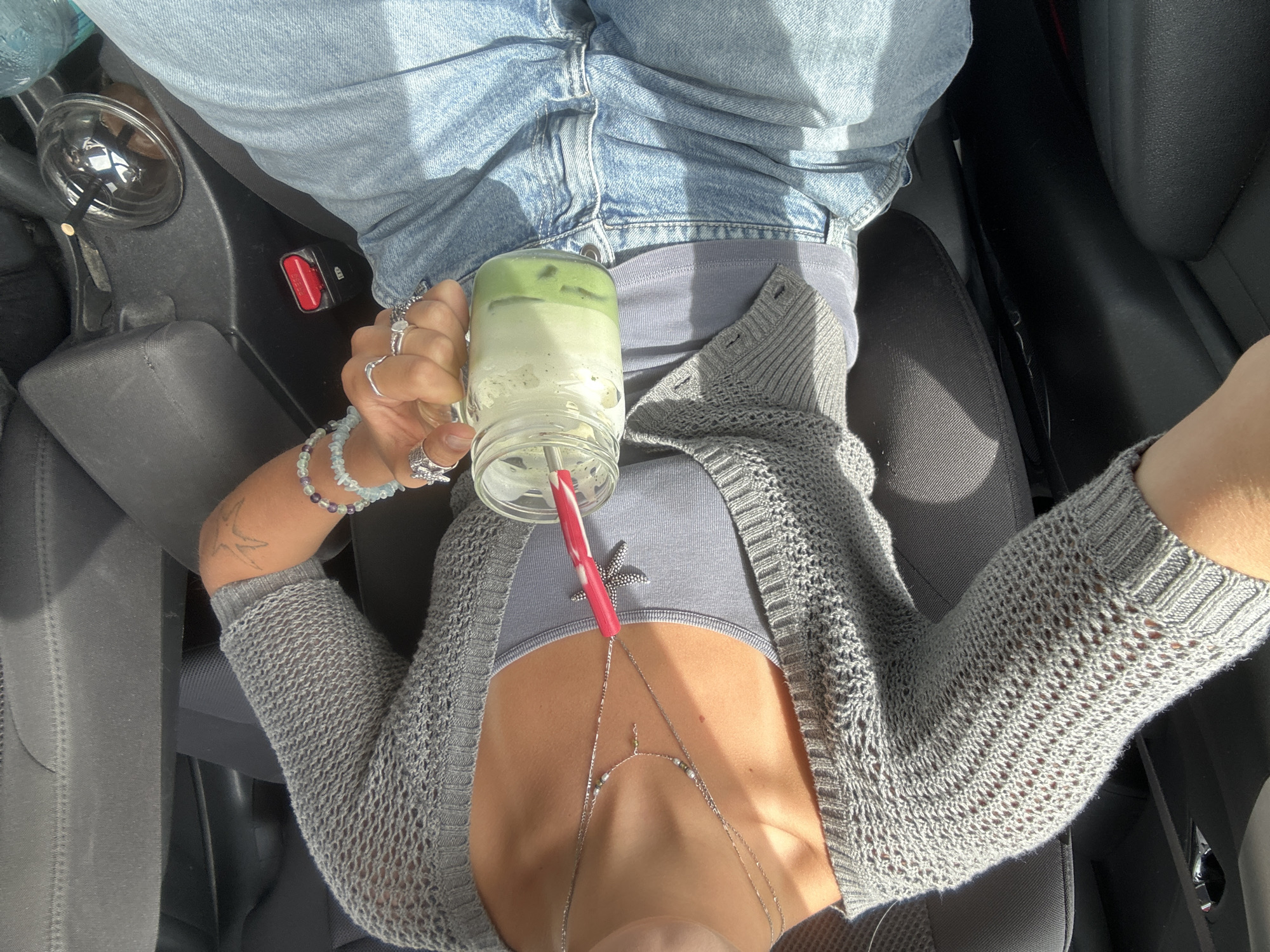 The image shows a person sitting in the driver's seat of a car, holding a mason jar with a green beverage and a red and white straw. The person is wearing a grey knitted cardigan, a grey tank top, and light blue denim jeans. They have several rings on their fingers and a beaded bracelet on their left wrist. A tattoo is visible on their left arm. The car's interior is partially visible, including a dark seat and part of the center console. A cup holder with a clear plastic cup and a straw is also visible. The lighting suggests it is daytime, with sunlight illuminating the scene. The location is not specified as Whistler, Canada, nor is there any visual evidence to support this claim.