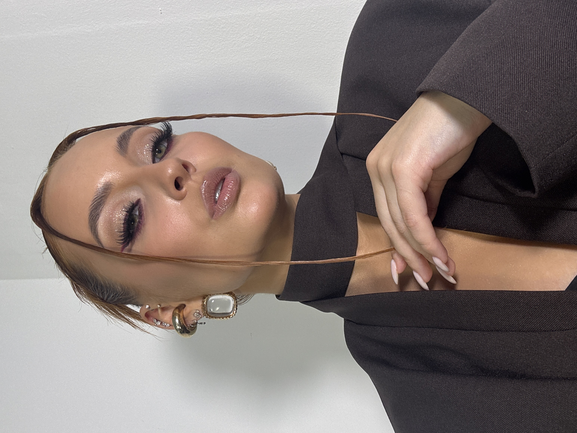 A female individual is depicted in a medium close-up portrait against a plain, light grey wall in an indoor environment. The specific location within Chaikovsky, Russia, is not discernible from the image. The subject has a light to medium skin tone, with full facial makeup including glittery eyeshadow, long dark false eyelashes, winged eyeliner, and glossy light pink lips. Her hair is pulled back, with two distinct, wet-look strands framing her face and extending downwards. Her visible left ear is adorned with multiple piercings: a large gold hoop, several small clear stud earrings, and a rectangular dangling earring featuring a milky white stone set in a gold frame. She is wearing a dark brown blazer with a structured, high-neck collar. Her right hand, with long, light pink manicured nails, rests gently on the left shoulder area of her blazer. The individual is looking directly at the viewer.