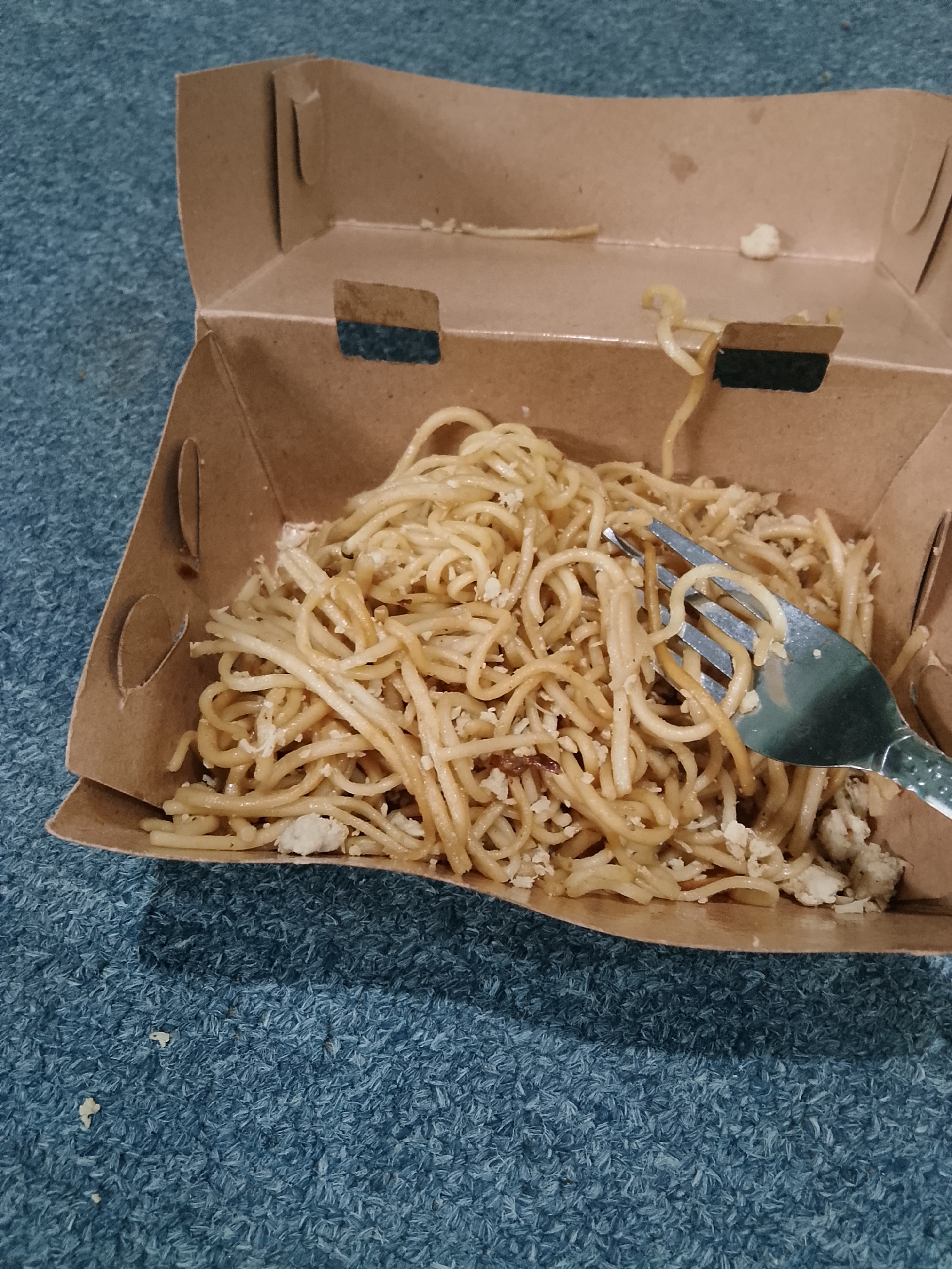 A close-up, overhead shot displays a portion of a paper takeout box filled with noodles on a textured, blue carpet. The box, made of corrugated cardboard, is open, with its sides angled upwards. The noodles, a light brown color, are tossed with what appears to be small pieces of white, possibly chicken or tofu, and a light brown sauce. A silver fork with a decorative pattern on its handle is partially submerged in the noodles, with a strand of noodles wrapped around one of its tines. The carpet has a high pile and is a deep blue with lighter blue and grey specks, suggesting it is outdoors, possibly on a street or in a park. No people are visible in the image. The overall impression is a casual, on-the-go meal. The provided metadata specifies the location as Tawangsari, Indonesia.