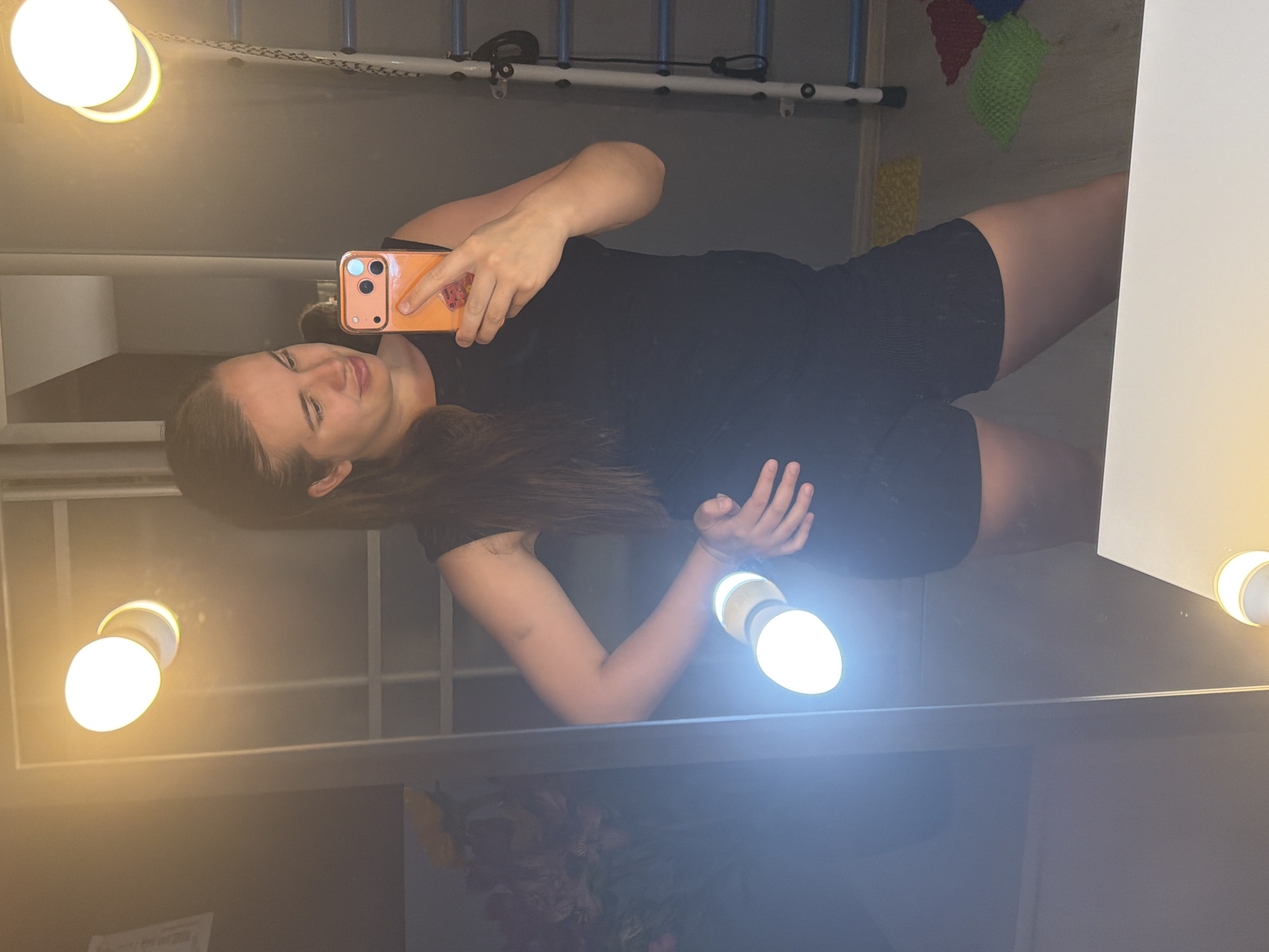 This is a mirror selfie, rotated 90 degrees clockwise, depicting a young woman taking a photograph with her smartphone. The woman, with long, dark hair, is visible from the waist up, wearing a dark (likely black) top and shorts or a romper. She holds an orange or peach-colored smartphone with a visible multi-lens camera array in her right hand, looking towards its screen with a slight, closed-mouth smile or smirk.
The scene appears to be an indoor setting, likely in front of a vanity mirror or a brightly lit area, given the presence of multiple exposed, glowing light bulbs. Two prominent bright white-yellow bulbs are visible on the left side of the frame, and another is partially seen on the right, casting significant glare and reflections across the image.
In the background, reflected in the mirror, various indoor elements can be discerned. Above the woman's head (in the rotated view), there's a structure with light-colored framing and blue horizontal bars, possibly part of a shelving unit or a bed frame. To the upper right, colorful knitted or woven items in red, green, and yellow are hanging against a wall. Towards the bottom of the frame, blurred floral patterns or other decorative objects are vaguely visible.
The artificial lighting makes the time of day undeterminable. No legible text is visible in the image. The location context for this image is Sochi, Russia.