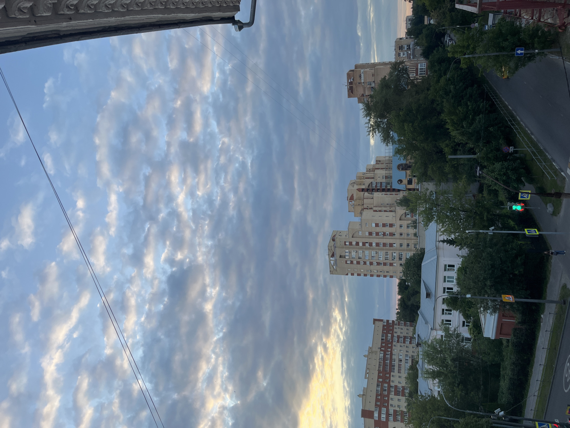 The image depicts an urban landscape at dusk in Ivanteyevka, Russia. The sky is filled with a mix of clouds, with patches of yellow and orange light suggesting the sun is setting or has recently set. Several multi-story apartment buildings, characteristic of Soviet-era architecture, are visible, some with a reddish-brick facade and others with a lighter, beige tone. Green trees and foliage are interspersed between the buildings and line the streets.
A road with painted lanes and traffic markings runs through the scene, with streetlights and traffic signals present. A few vehicles are parked or driving on the road. There are a couple of pedestrians visible, one walking on the sidewalk and another further down the street. A balcony or architectural detail from a building is visible in the upper right corner, framing the scene. Overhead power lines traverse the sky. The overall impression is of a residential area in a Russian city during the evening.