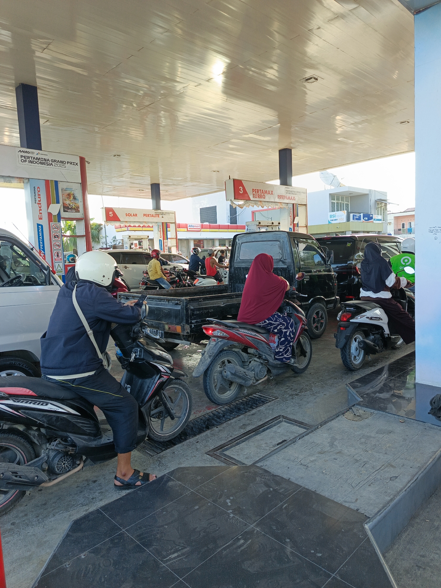 The image depicts a scene at a Pertamina gas station in Tanakawu, Indonesia. Several vehicles, including motorcycles and a pickup truck, are lined up at the pumps, suggesting a queue. People are seated on or near their motorcycles, some wearing helmets. One person in the foreground is looking at their phone while seated on a motorcycle. The gas station canopy is visible overhead, with signage indicating fuel types such as "Pertamax Turbo" and "Solar." In the background, buildings and a satellite dish are present, indicating a developed area within the city. The ground is paved with concrete and tiles.