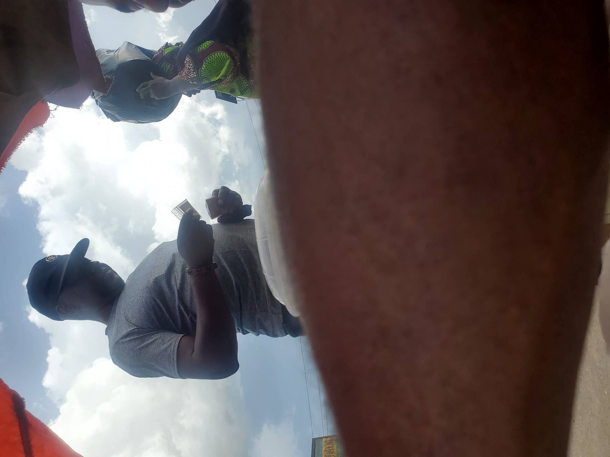 The image shows a low-angle shot of several people in what appears to be an outdoor market setting in Gwong, Nigeria. In the foreground, a large portion of a person's arm and leg fills the right side of the frame. To the left, a man wearing a grey t-shirt and a black baseball cap is visible from the chest up, looking to his left. He has a bracelet on his wrist. Behind him and slightly to the left, a person is holding a small, clear object, possibly a phone or a card. Further in the background, another person is wearing a colorful patterned top. The sky above is cloudy, suggesting it might be daytime. The overall impression is a candid snapshot of a moment in a busy public space.