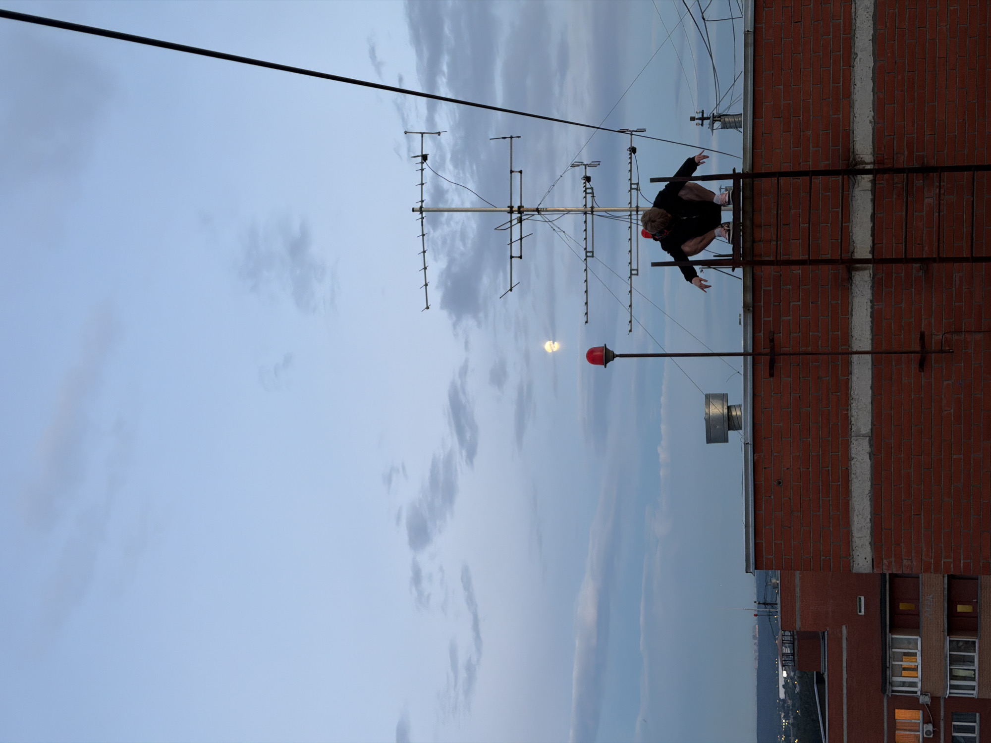 A person is crouched on the rooftop of a brick building, appearing to be performing a stunt or posing for a photo. They are wearing a black hoodie, black shorts, and red and white sneakers. The person has blond hair and is wearing goggles. They are positioned on a metal structure that looks like a ladder or scaffolding, with a red warning light visible below them. In the background, a large television antenna dominates the frame, with wires extending across the sky. The sky is filled with clouds, suggesting it might be evening or overcast. The image is taken from a low angle, looking up at the person and the antenna. The overall scene is set on a rooftop in Khimki, Russia, with glimpses of other buildings and a distant cityscape visible. The lighting suggests it is late afternoon or early evening. There is no visible text in the image.