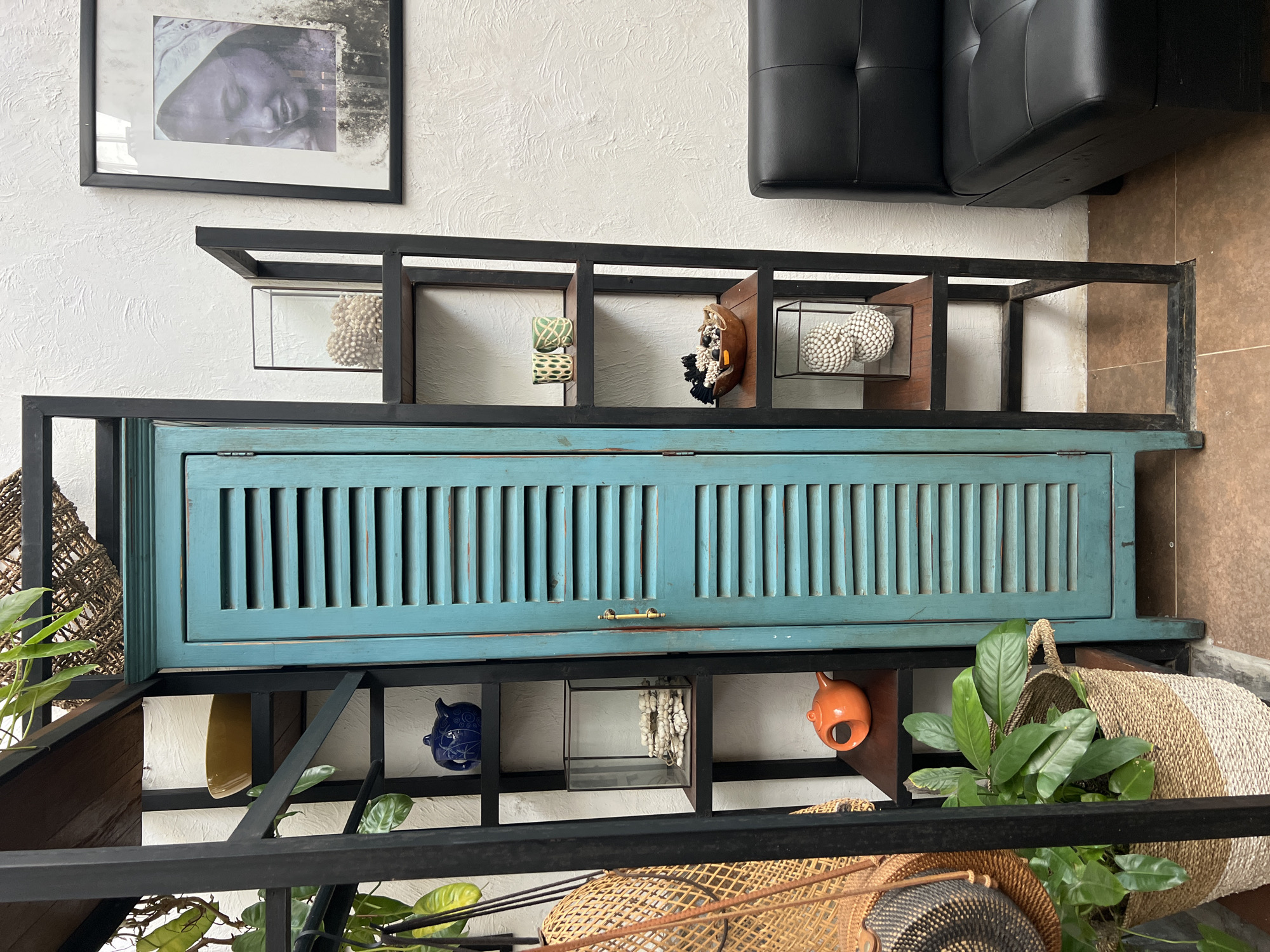 A medium shot captures a piece of furniture against a white textured wall. The piece features a cabinet with blue horizontal slats above and below a black metal frame. The frame has various compartments holding decorative items, including baskets and pottery. A framed print of a face hangs on the wall above the furniture. The scene is located in North Kuta, Indonesia. A black couch is also visible above the furniture.