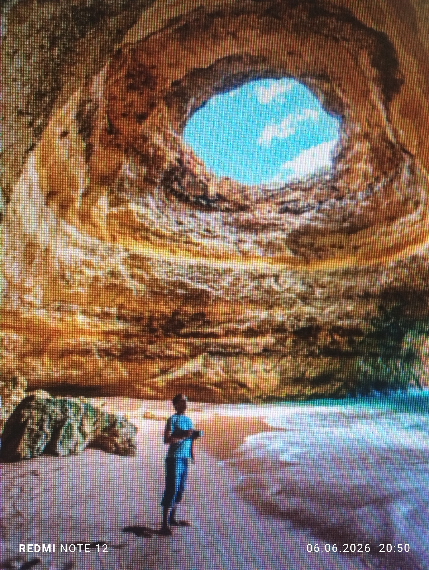 The image displays the interior of a large sea cave, characterized by a prominent circular opening in its roof that reveals a bright blue sky with white clouds. The cave walls are composed of stratified, earthy-toned rock formations, predominantly orange, brown, and ochre. A sandy beach occupies the cave floor, with ocean waves breaking on the right side, forming white foam. On the left side of the beach, some darker rock formations are visible.
A lone individual, an adult, stands near the center-right of the beach, facing towards the ocean. They are dressed in a light-colored top and darker lower garments, holding a small object in their hands. A shadow is cast from the person onto the sand. The image exhibits a distinct pixel grid pattern, suggesting it is a photograph of a digital screen, and possesses a moderate level of blur. Digital watermarks at the bottom corners read "REDMI NOTE 12" on the left and "06.06.2026 20:50" on the right. This scene is described as being located in the city of Burgas, Bulgaria.