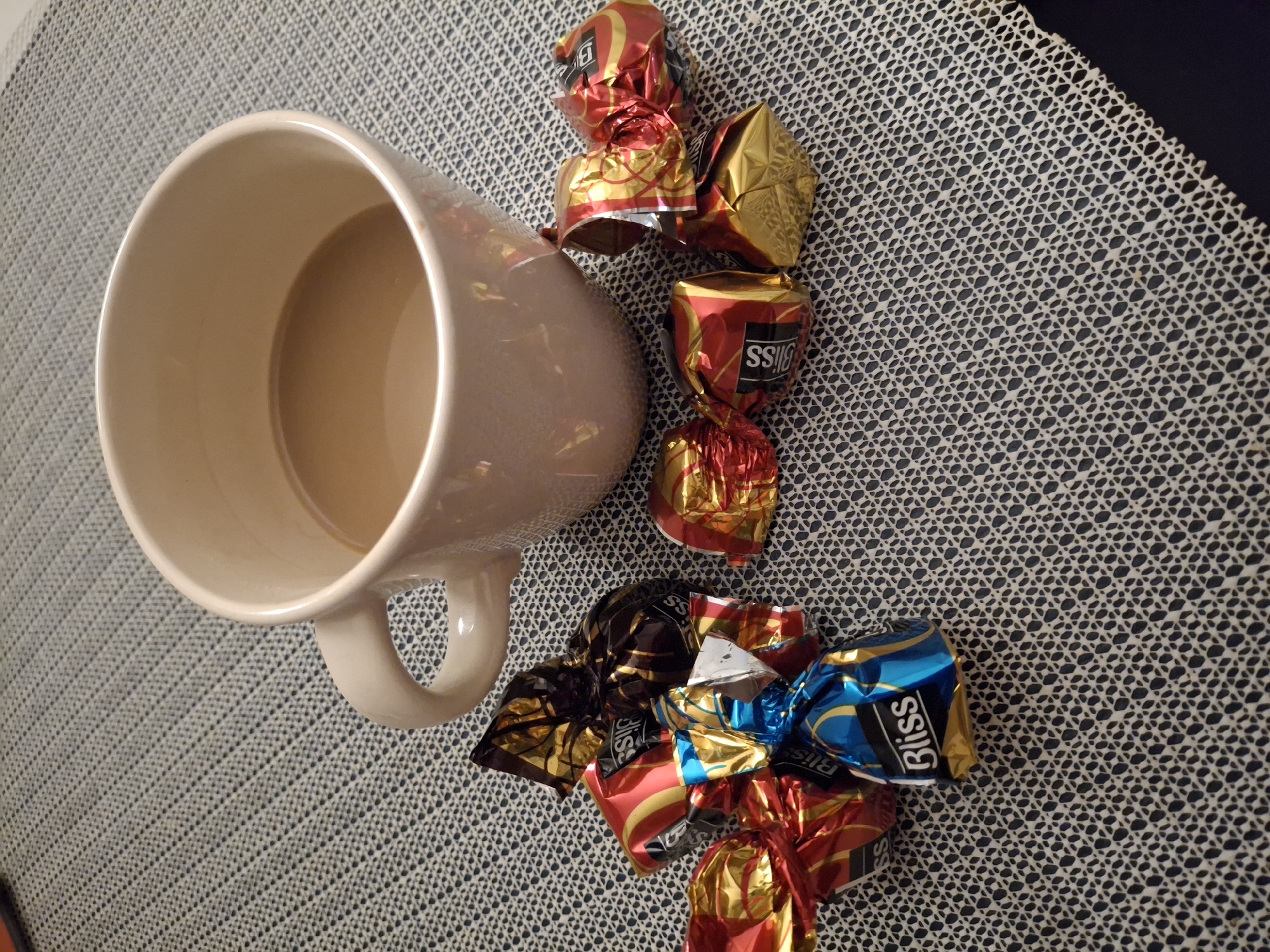 The image shows a beige ceramic mug partially filled with a light brown liquid (likely coffee or tea with milk) placed on a patterned grey and white placemat. To the right of the mug, there are several shiny, wrapped chocolates with the word "Bliss" visible on some of the wrappers. The wrappers are in a variety of colors, including red, gold, blue, and brown. The placemat has a textured, woven pattern. The background is blurred, but a dark surface is visible in the upper right corner. The location is not discernible based on the image and cannot be assumed to be Eemshaven, Netherlands.