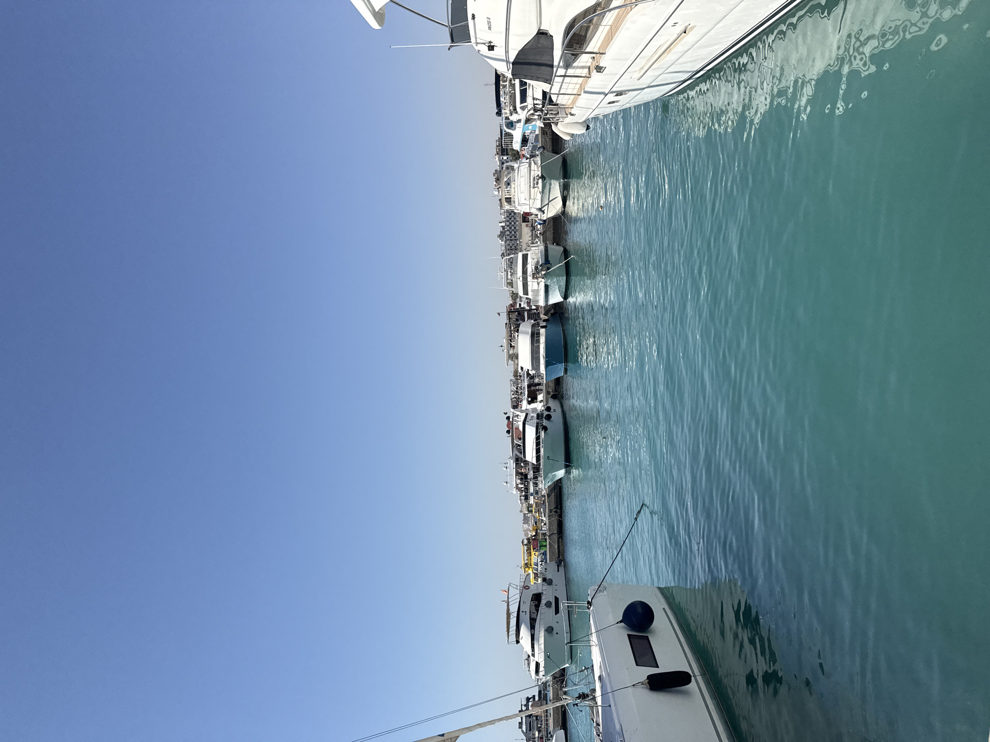 A wide shot captures a serene view of a marina in Hurghada, Egypt, under a clear, bright blue sky. The foreground is dominated by the turquoise waters of the sea, gently rippling. On the left, the stern of a white sailboat is visible, with its hull partially submerged, and dark blue fenders hanging off its side. A rope extends from the sailboat into the water. In the mid-ground, a line of various white and blue motor yachts and smaller boats are moored along a dock. These vessels vary in size and design, with some featuring multiple decks and awnings. The boats are neatly arranged, suggesting an organized and well-maintained marina. The background reveals a strip of buildings, some appearing to be hotels or resorts, lining the shore. The overall atmosphere is peaceful and sunny, indicative of a pleasant day at the seaside.