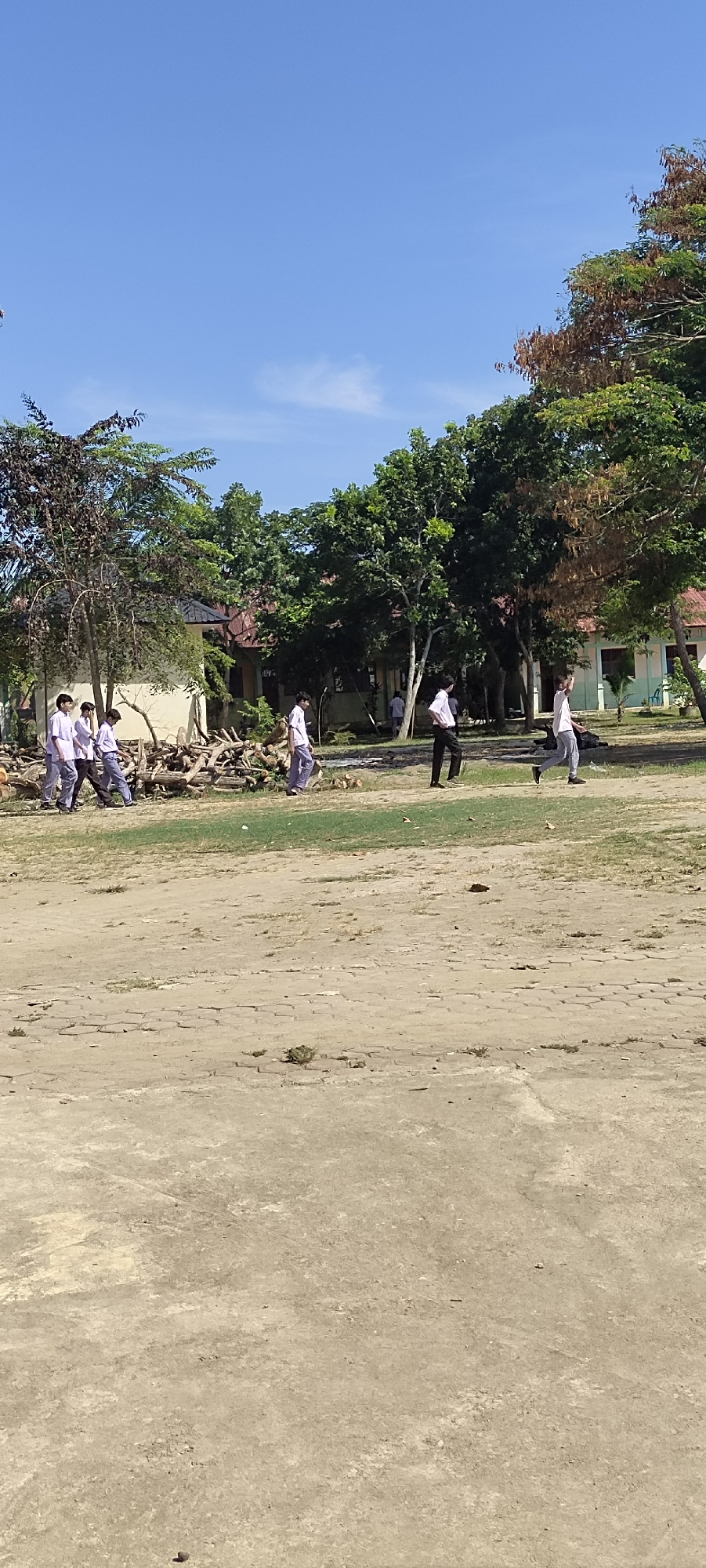 The image depicts an outdoor scene under a clear blue sky, likely in Langnibong, Indonesia. In the foreground, a sandy, cracked ground surface dominates the lower half of the frame. The middle ground shows a patch of dry grass and further back, a large pile of cut logs and branches. Beyond the logs, a school building with red-tiled roofs is visible, partially obscured by lush green trees and some trees with brown foliage. Several young people, appearing to be students in uniform, are walking across the grassy area. Some are walking in a group of four on the left, wearing white shirts and dark pants. Another individual in similar attire is seen walking alone in the middle of the frame, and two more individuals in white shirts and dark pants are visible further to the right. The overall impression is of a school environment during daylight hours.